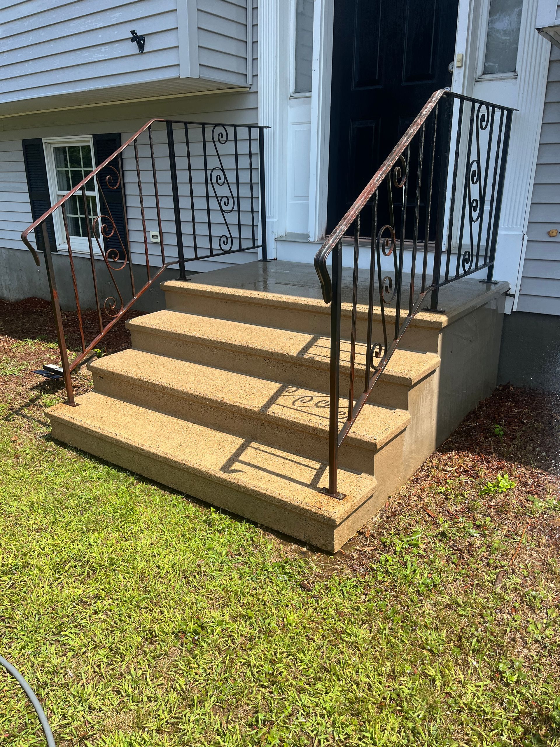 Front porch steps with tan concrete stairs and black railings beside a house, bordered by grass.