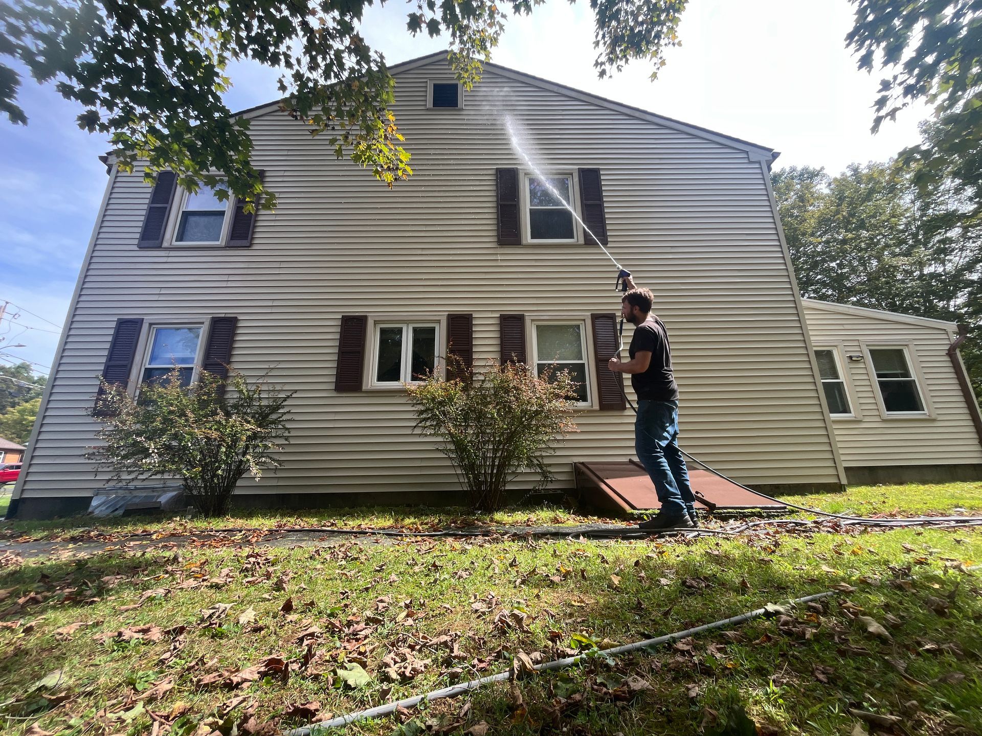Person spraying water at the side of a beige house in a sunny yard