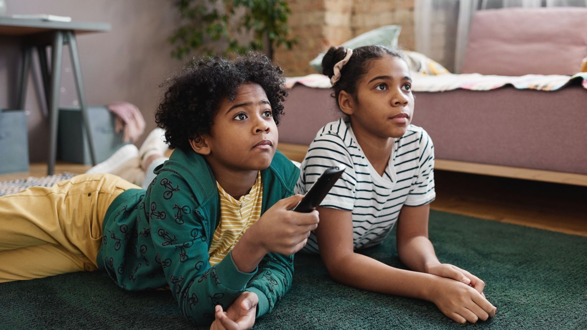 Two children lying on a carpet, watching TV and holding a remote control