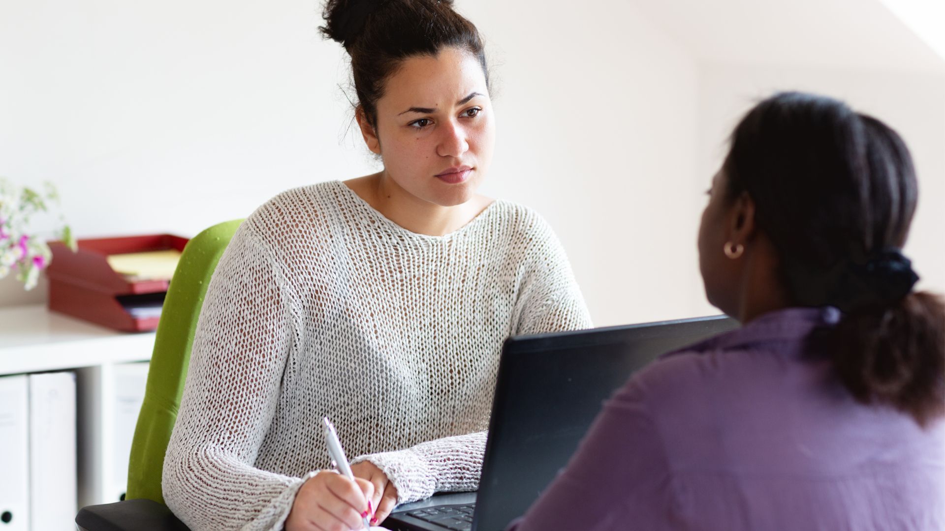 Two people talking at a table with a laptop and notebook in a bright office setting