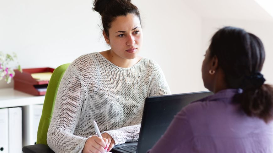 Two people talking at a table with a laptop and notebook in a bright office setting
