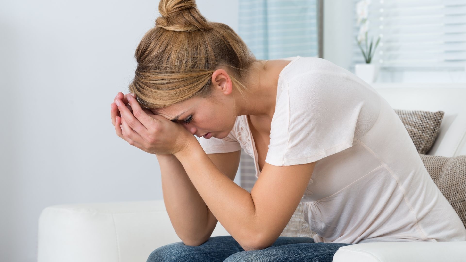 Woman sitting on a couch with head in hands, looking upset in a bright living room.