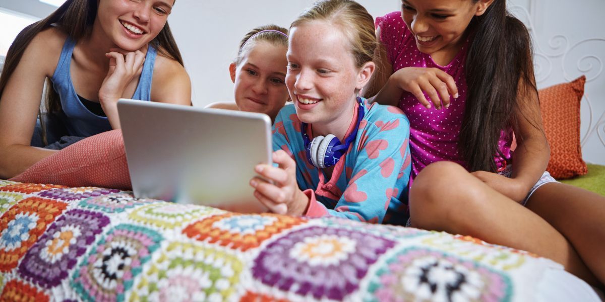 Four people, smiling, look at a tablet together on a bed with a colorful blanket.