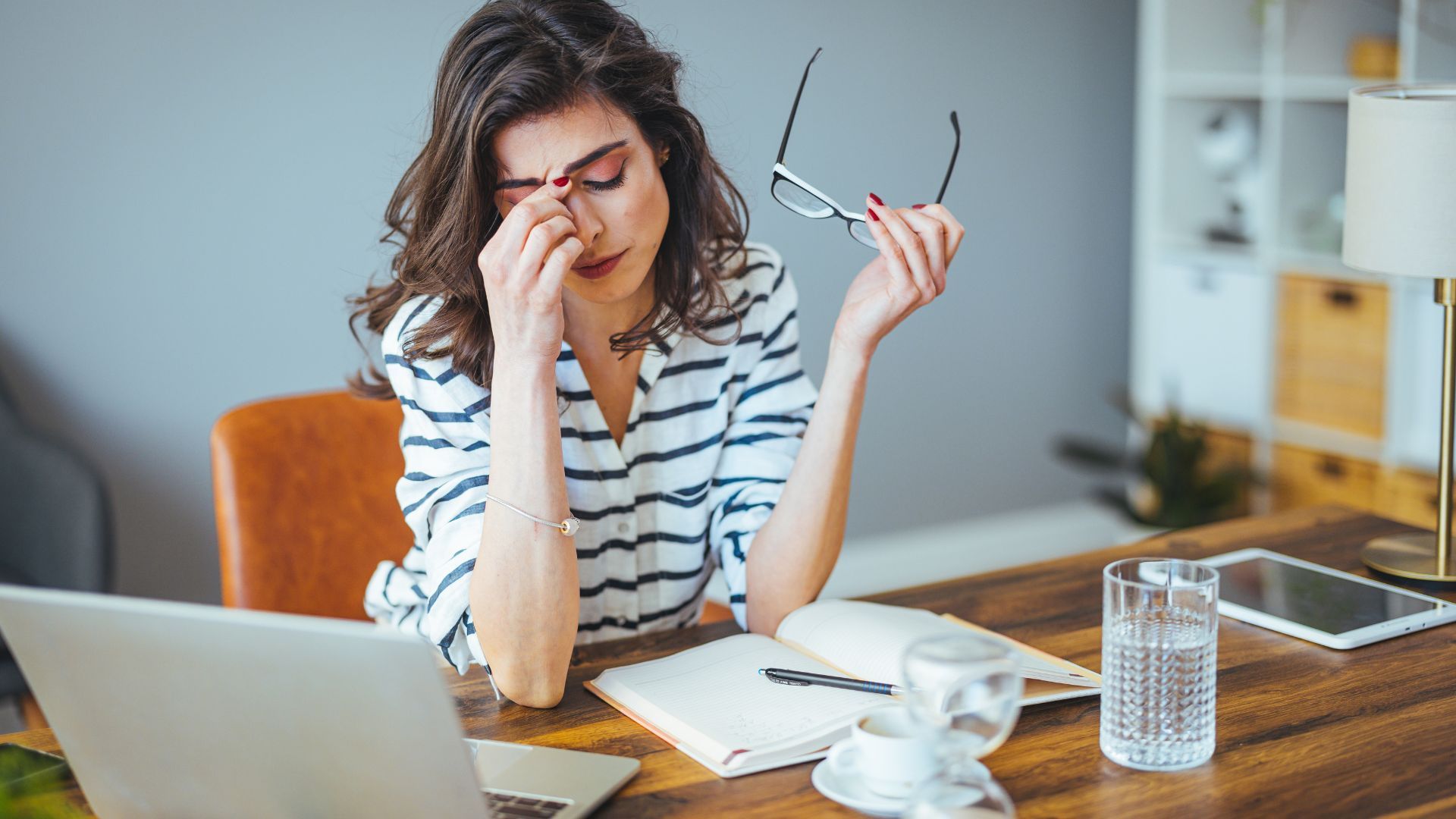 Stressed person at a desk rubbing their eyes, holding glasses, with a laptop, papers, and coffee mug nearby