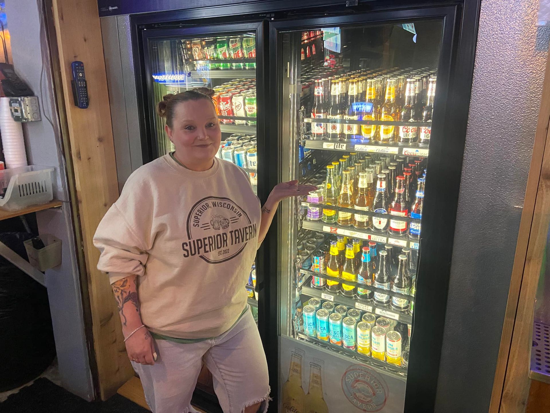 Woman with tattoos points to a cooler stocked with drinks in a store.