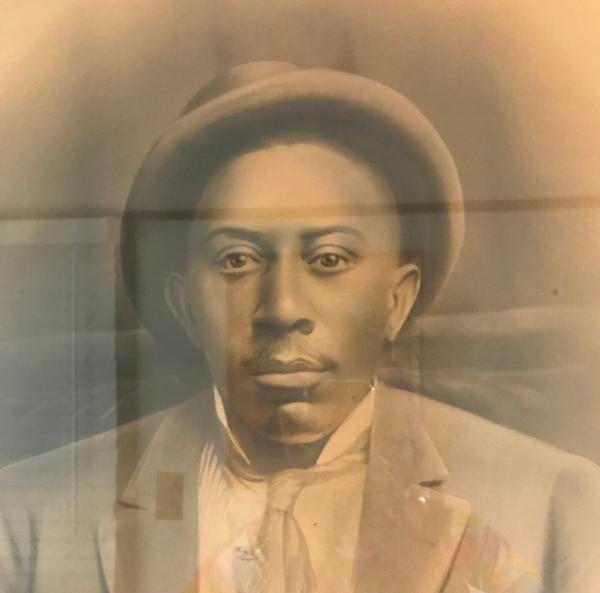 Sepia-toned portrait of a person in a hat and suit, facing forward with a calm expression.