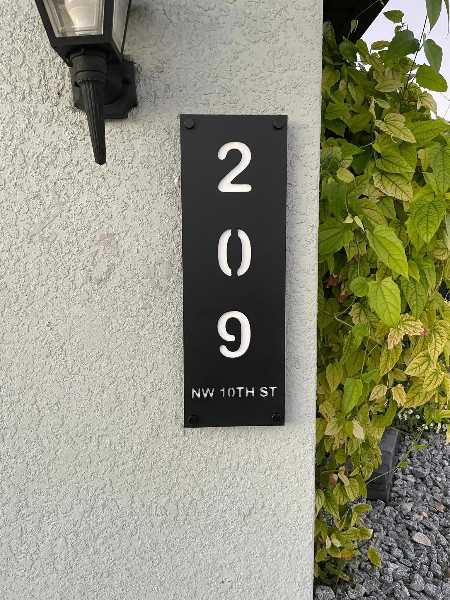 Black house number sign on a light-colored wall, with white numbers and street name; a black lamp above.
