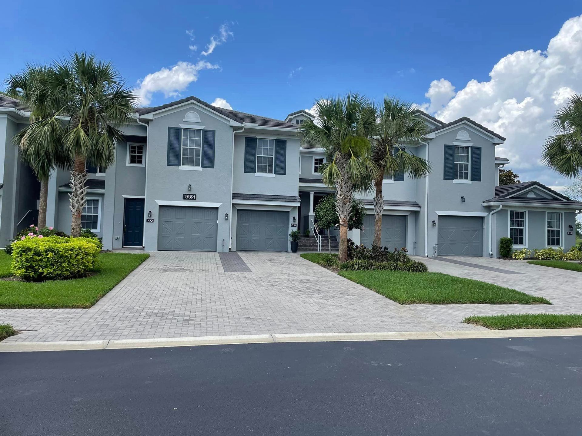 Row of blue-gray townhouses with gray garage doors, palm trees, and brick paved driveways under a blue sky.