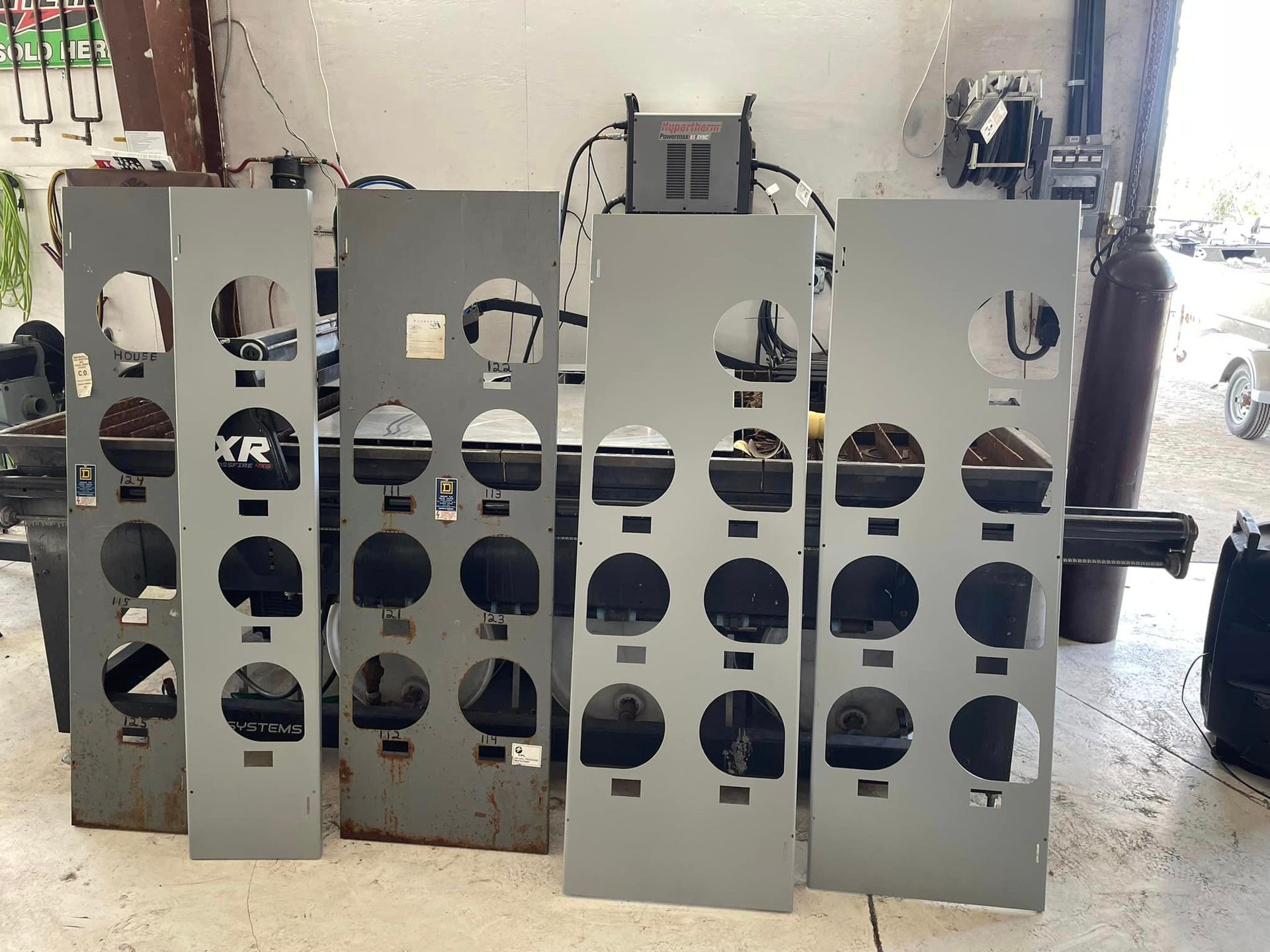 Five gray metal panels with various round and square cutouts, likely for machinery, in a workshop.