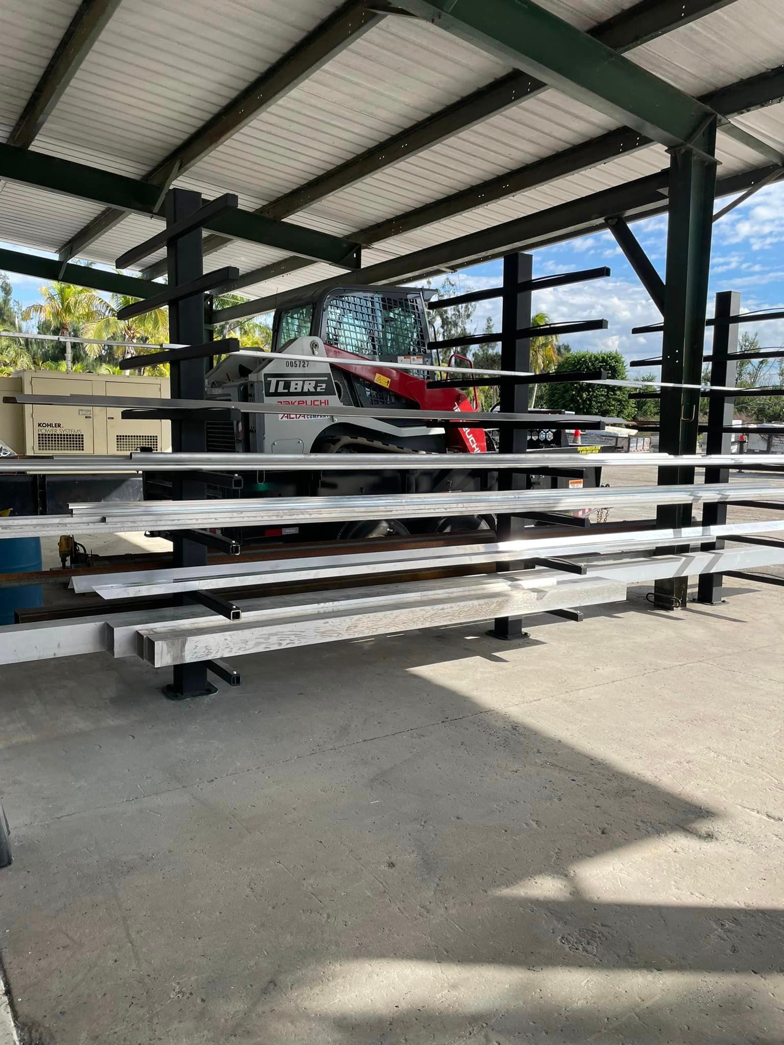 Steel beams stored on an open-air rack, with a Bobcat skid steer in the background, outdoors.