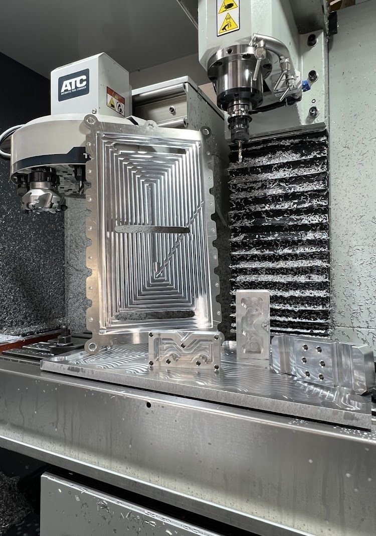 A CNC machine milling a rectangular metal part with intricate interior grooves. Coolant and chips are present.