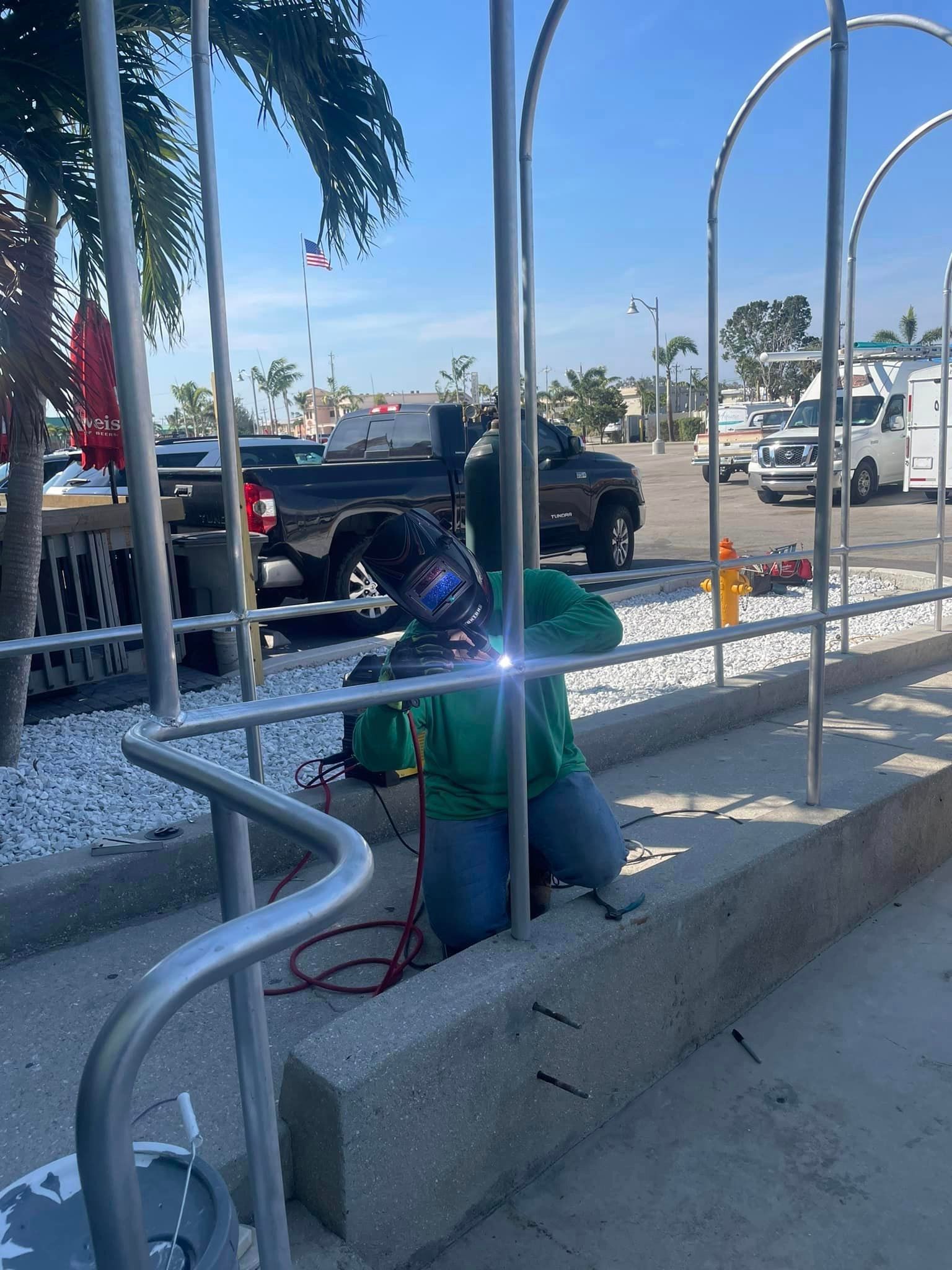 Welder in green shirt welds metal railing outdoors on a sunny day.