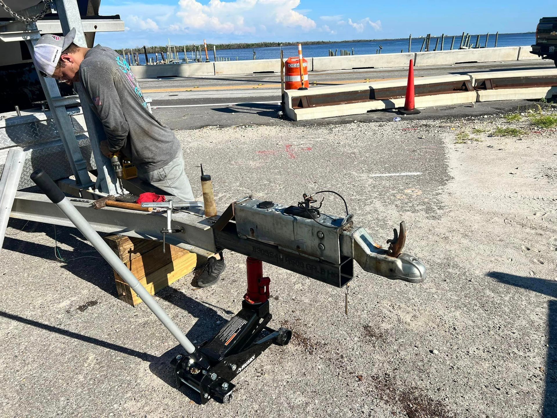 Man working on a boat trailer outdoors, using a jack. Gray metal, gray concrete, blue water in the background.