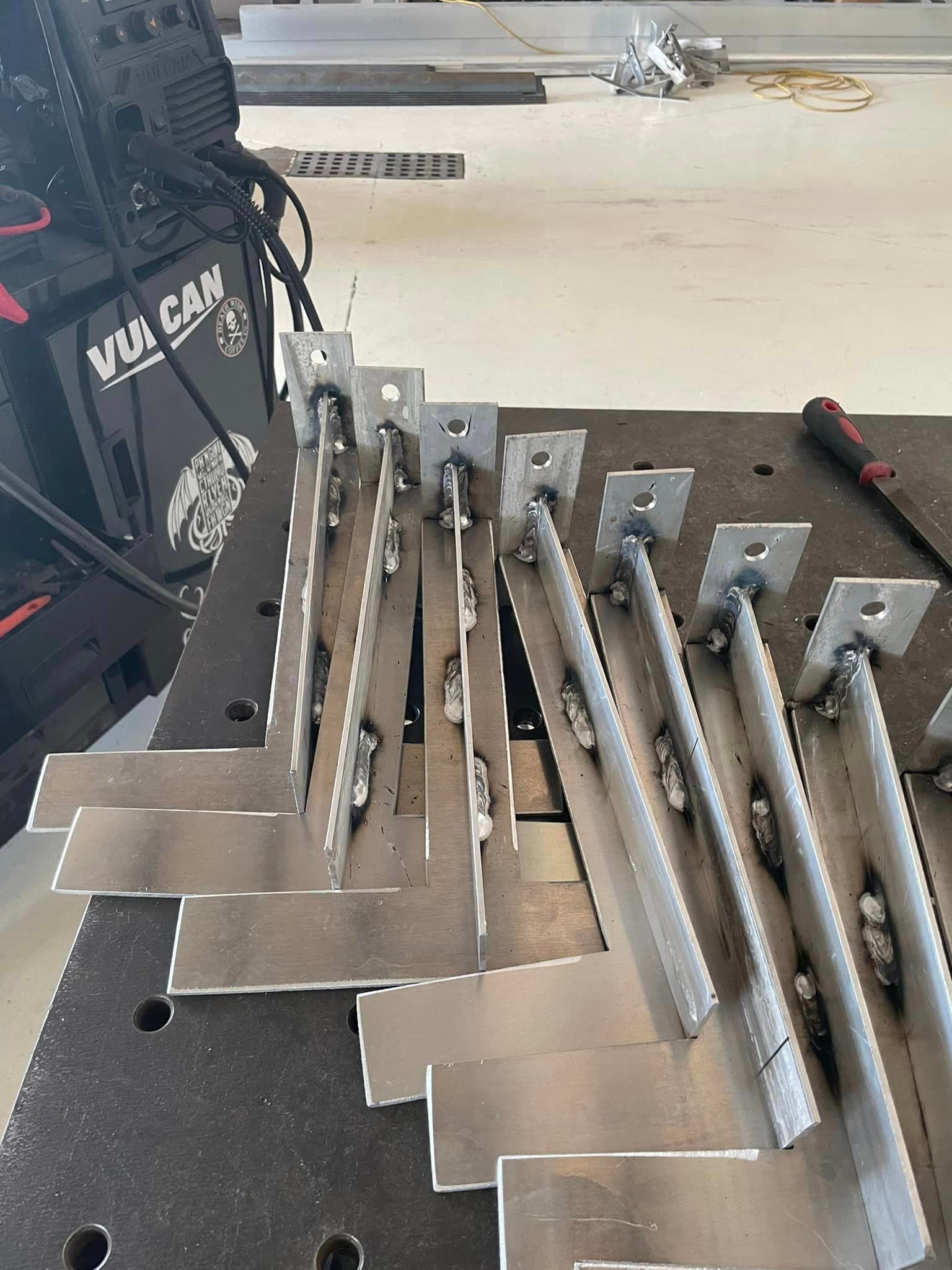 Metal brackets welded on a metal table in a workshop. A welding machine is visible in the background.
