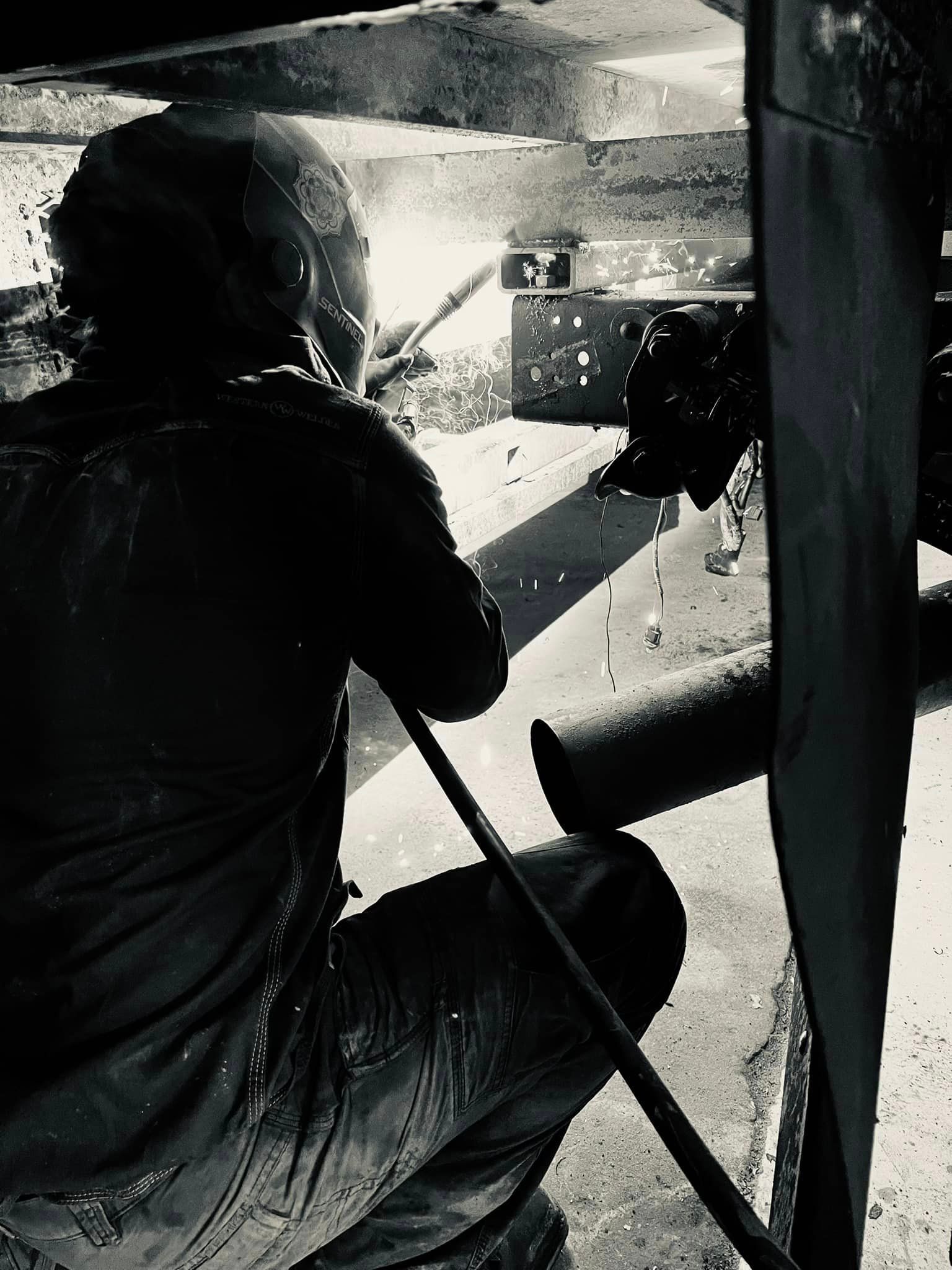A welder working under a structure, sparks flying. Black and white photo.