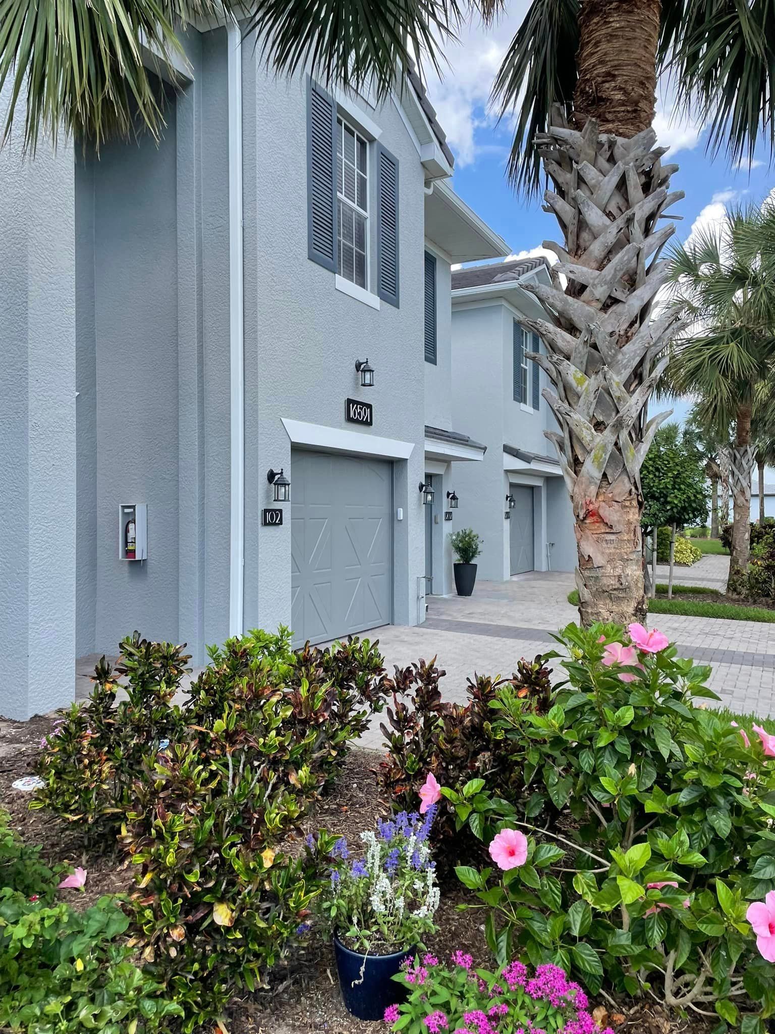 Two-story light blue townhouses with garages, flanked by tropical landscaping, palm trees, and a brick walkway.