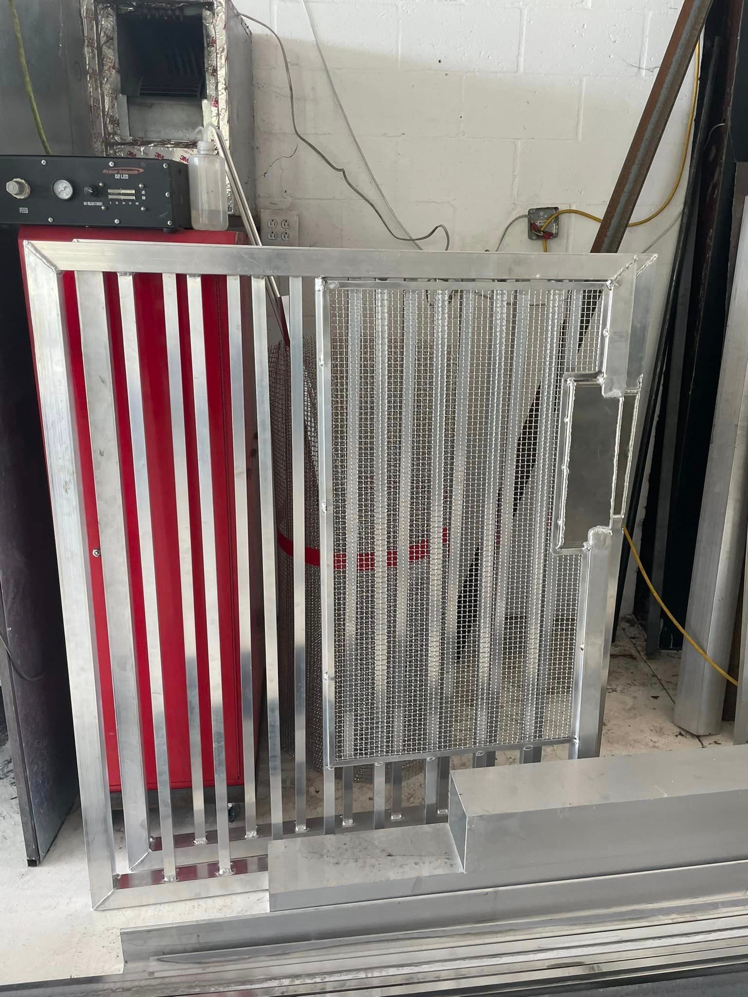 A metal filter with vertical bars and a mesh, leaning against a red wall, with other metal pieces.