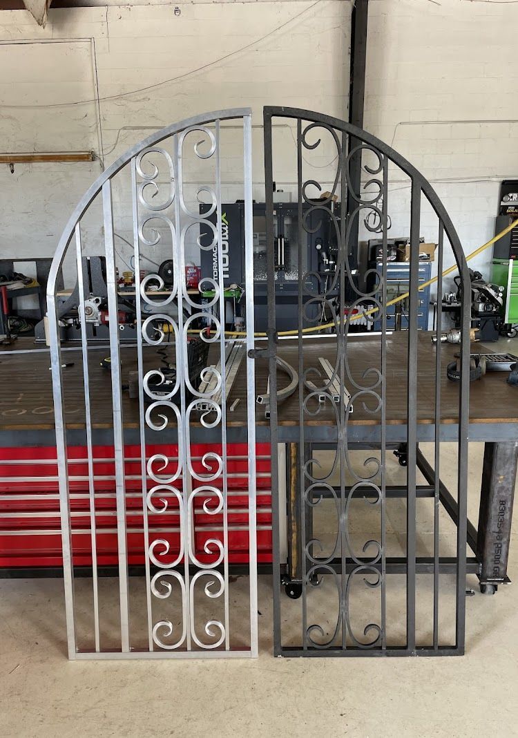 Two metal gates, one silver and one gray, stand side-by-side in a workshop setting.