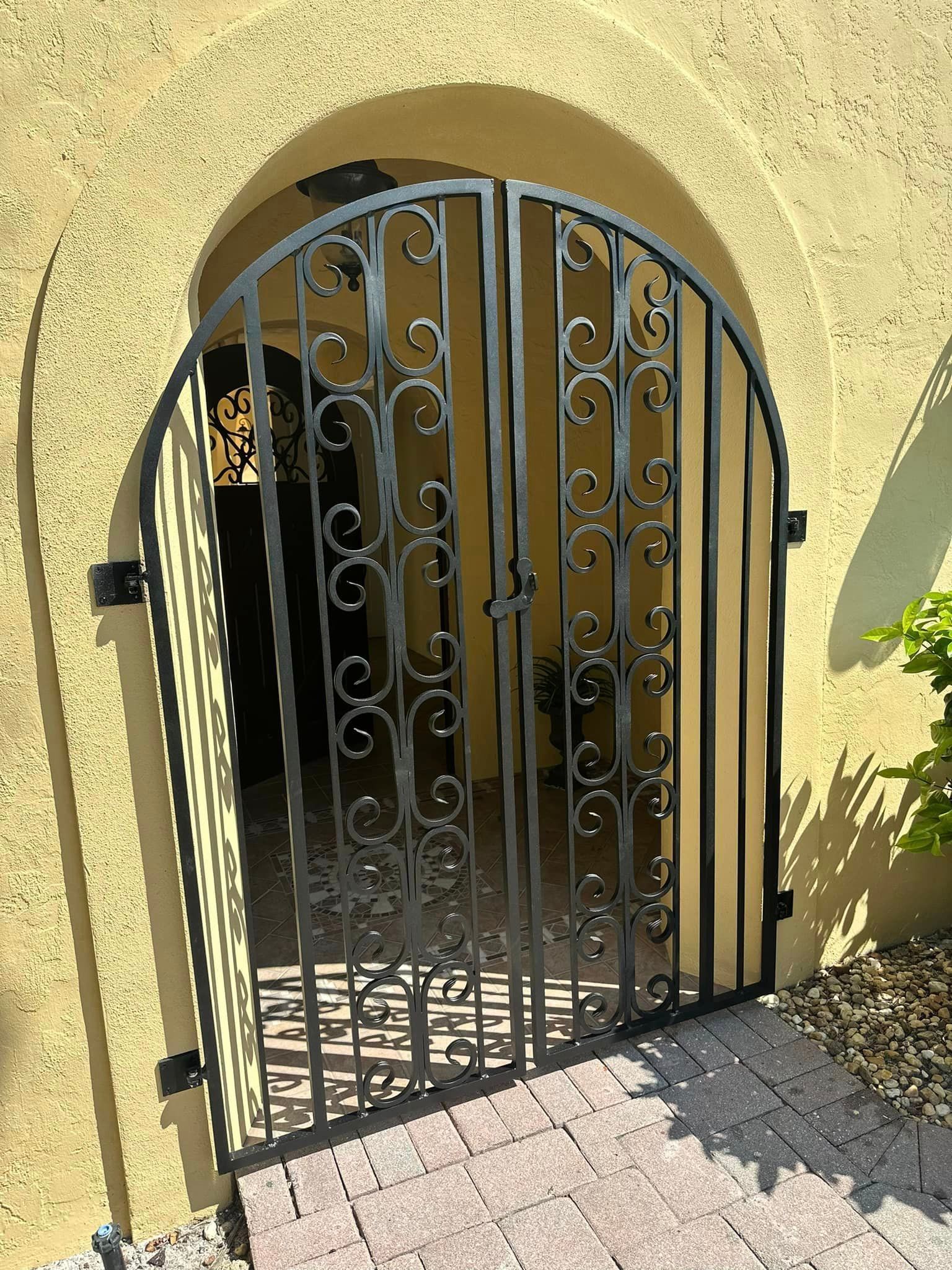 Black wrought-iron gate, open slightly, set in a yellow arched entryway, leading to a darker interior.