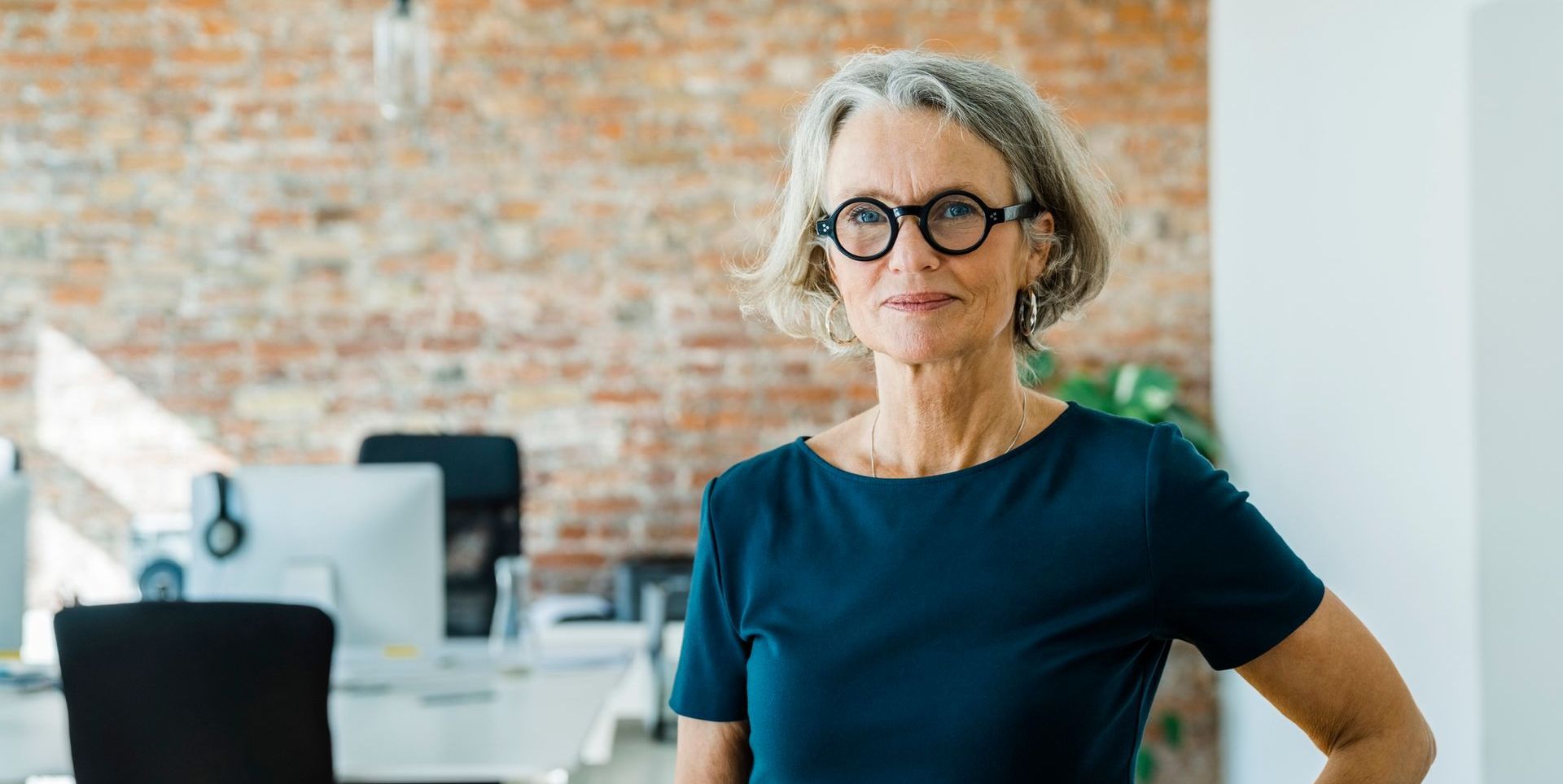 A woman wearing glasses is standing in an office with her hands on her hips.