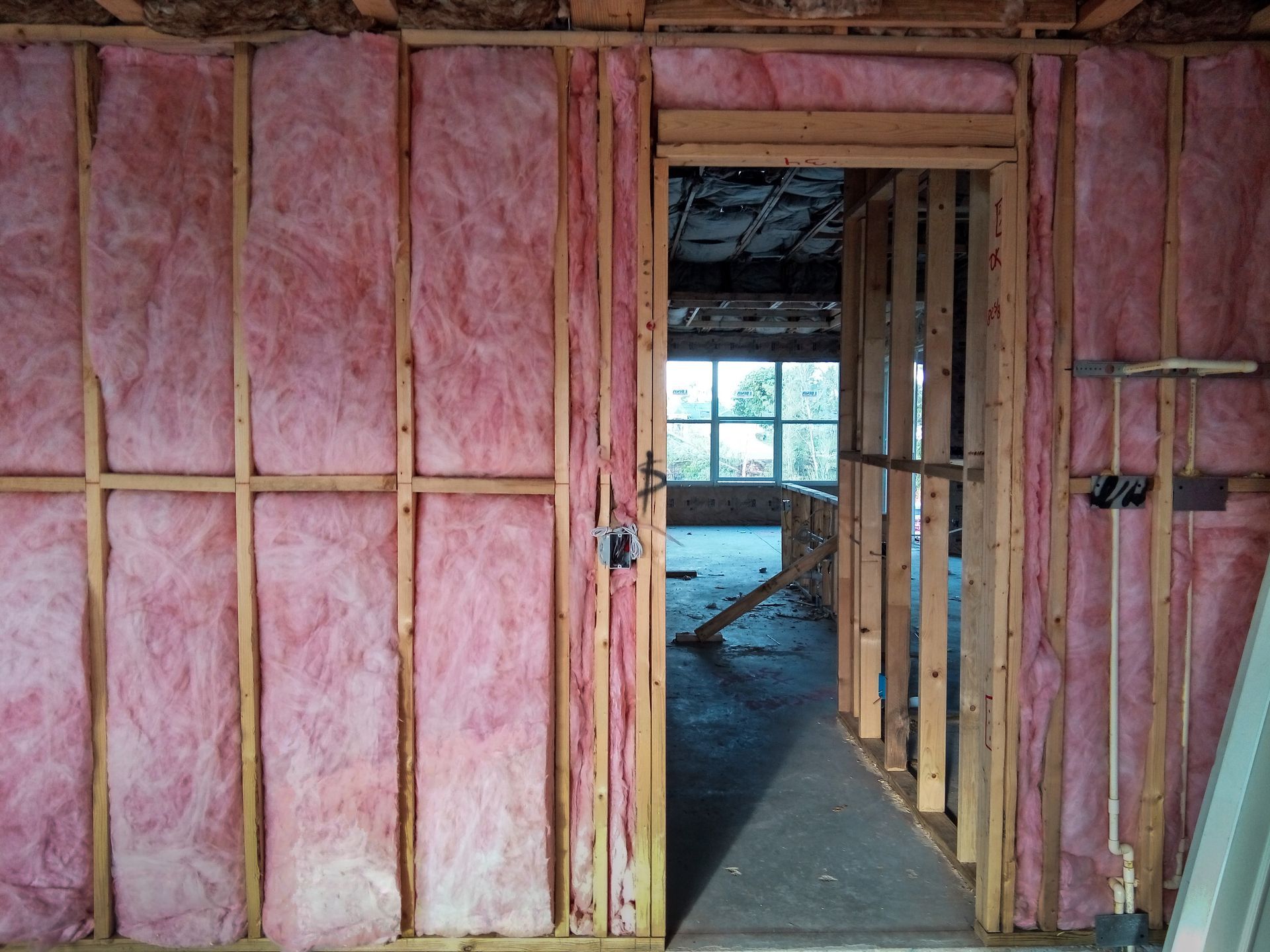 Interior wall framing with pink insulation and an open doorway into a room with windows.
