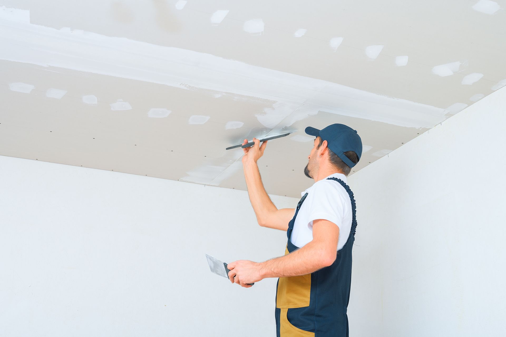 Worker sanding a ceiling patch with a putty knife in a white room