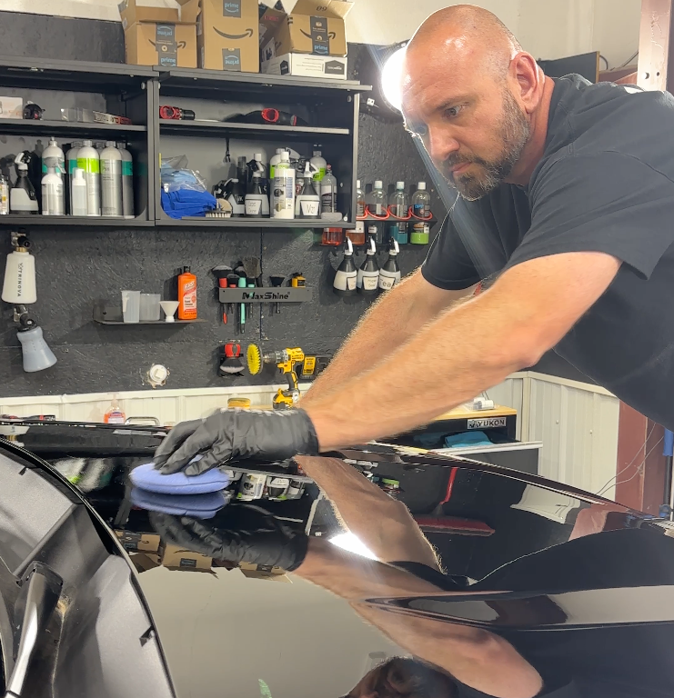 Man polishing a black car hood in a garage, with shelves of cleaning supplies in the background