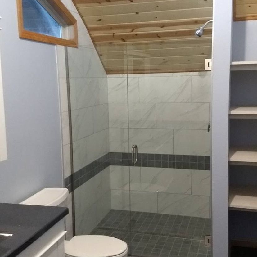Bathroom with a glass shower, toilet, and shelving; gray and white tile, wooden ceiling.