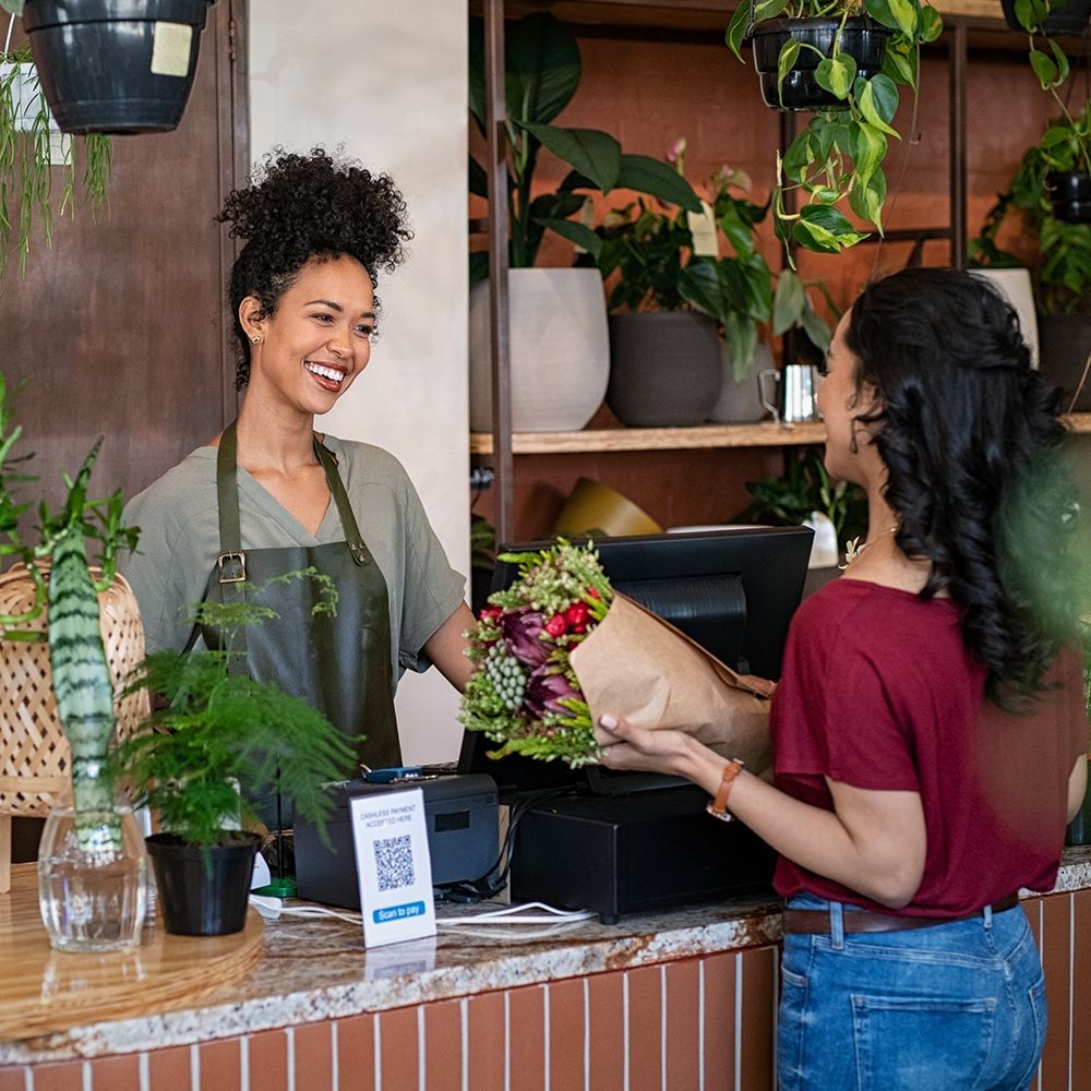 Florist smiling at counter, handing a bouquet to a customer in a plant-filled shop
