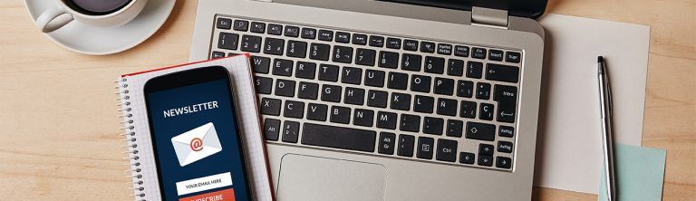 A laptop , cell phone , notebook and cup of coffee on a wooden table.
