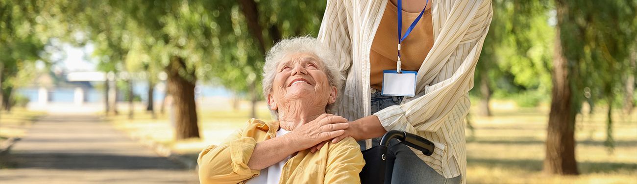 A woman is standing next to an elderly woman in a wheelchair in a park.