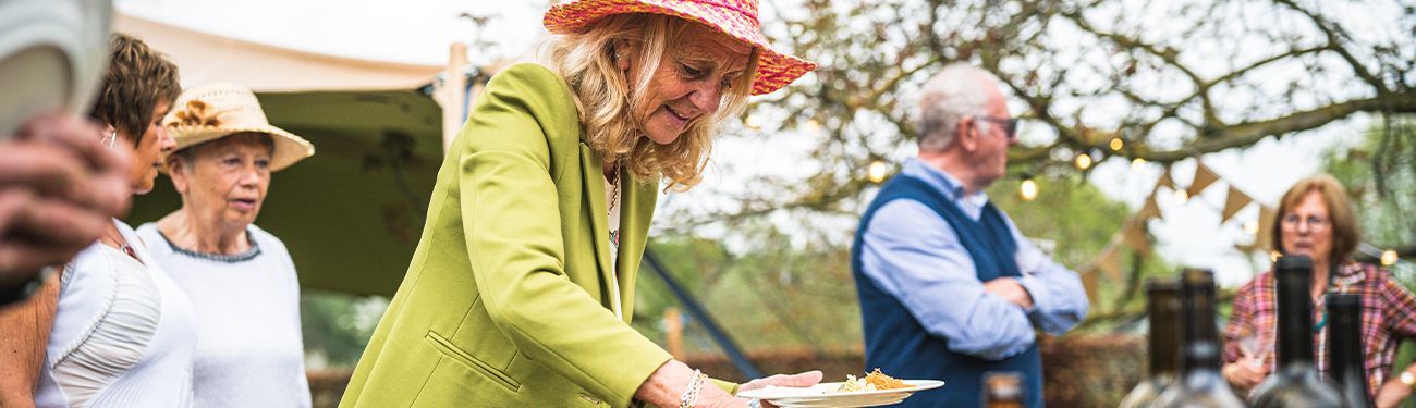 People outdoors at a garden event, with a woman in a green coat serving food at a table.