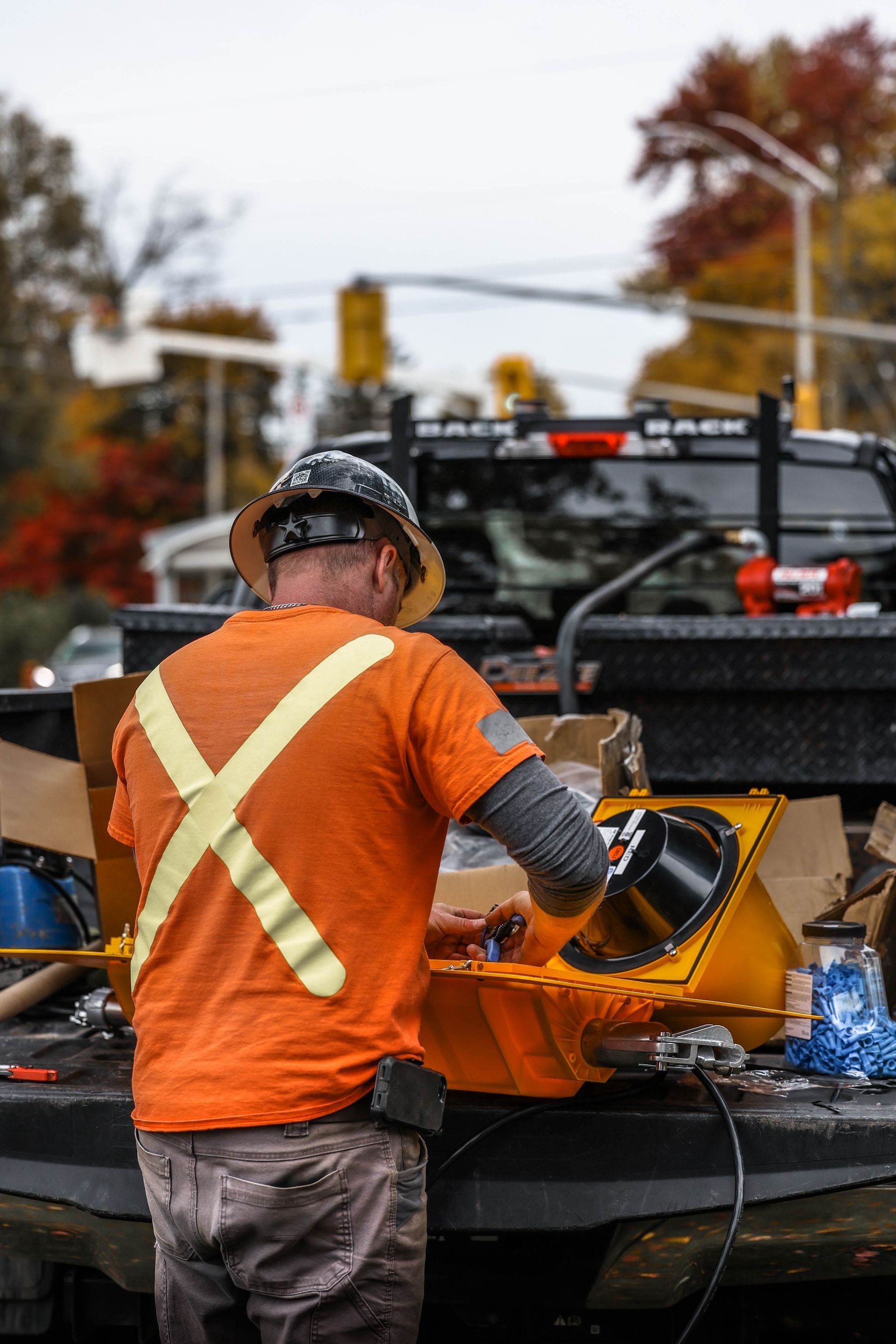 Construction worker in orange safety shirt with X-back, working on equipment on truck bed, blurred street background.