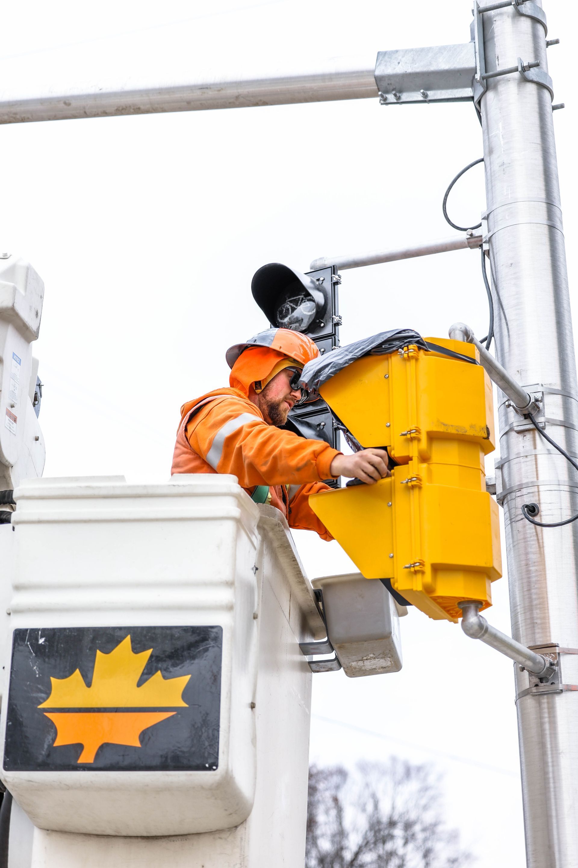 Worker in orange safety gear in a lift bucket, repairing a yellow traffic light.