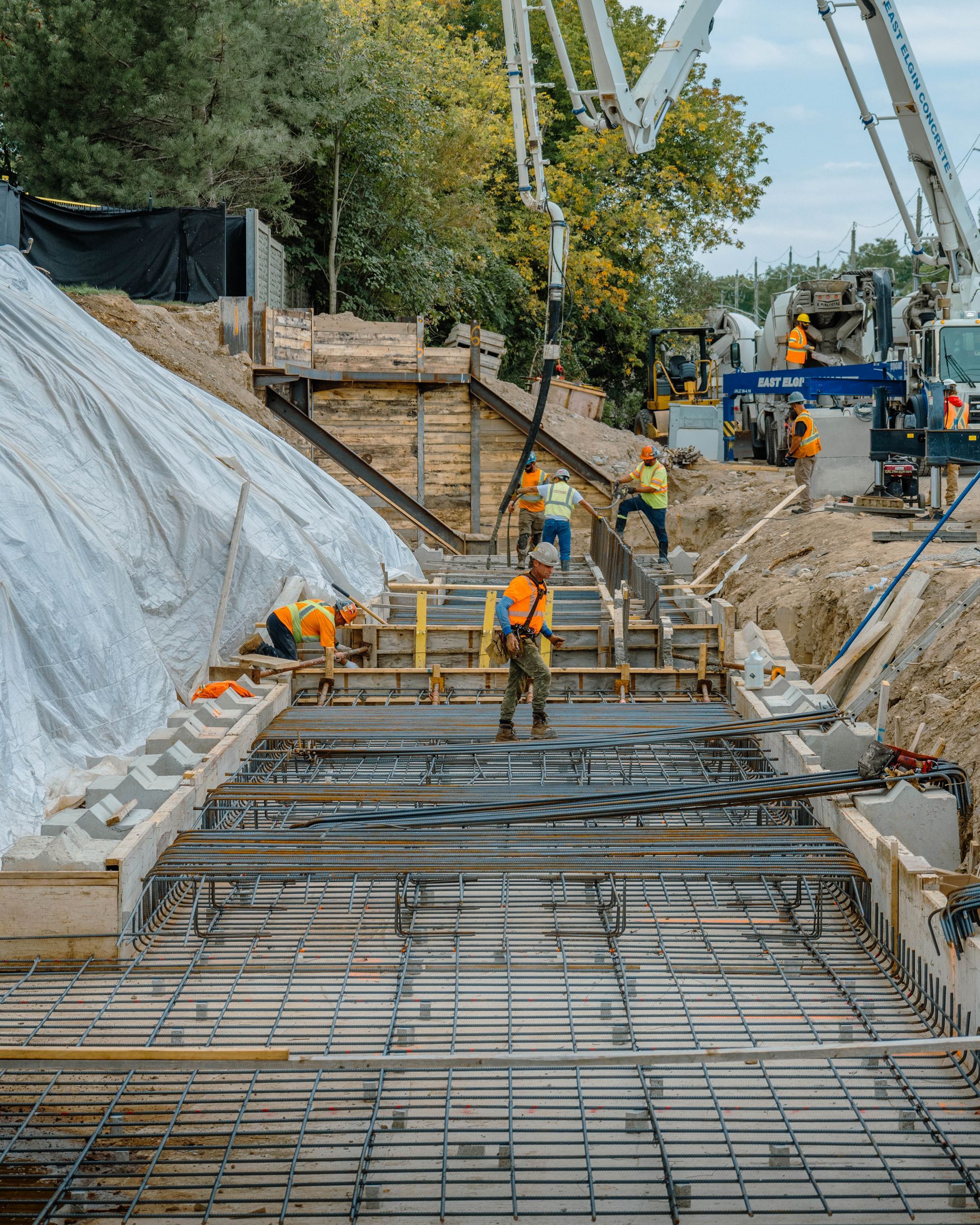 Construction workers pouring concrete at a site with a concrete pump. Rebar and wooden forms are visible.