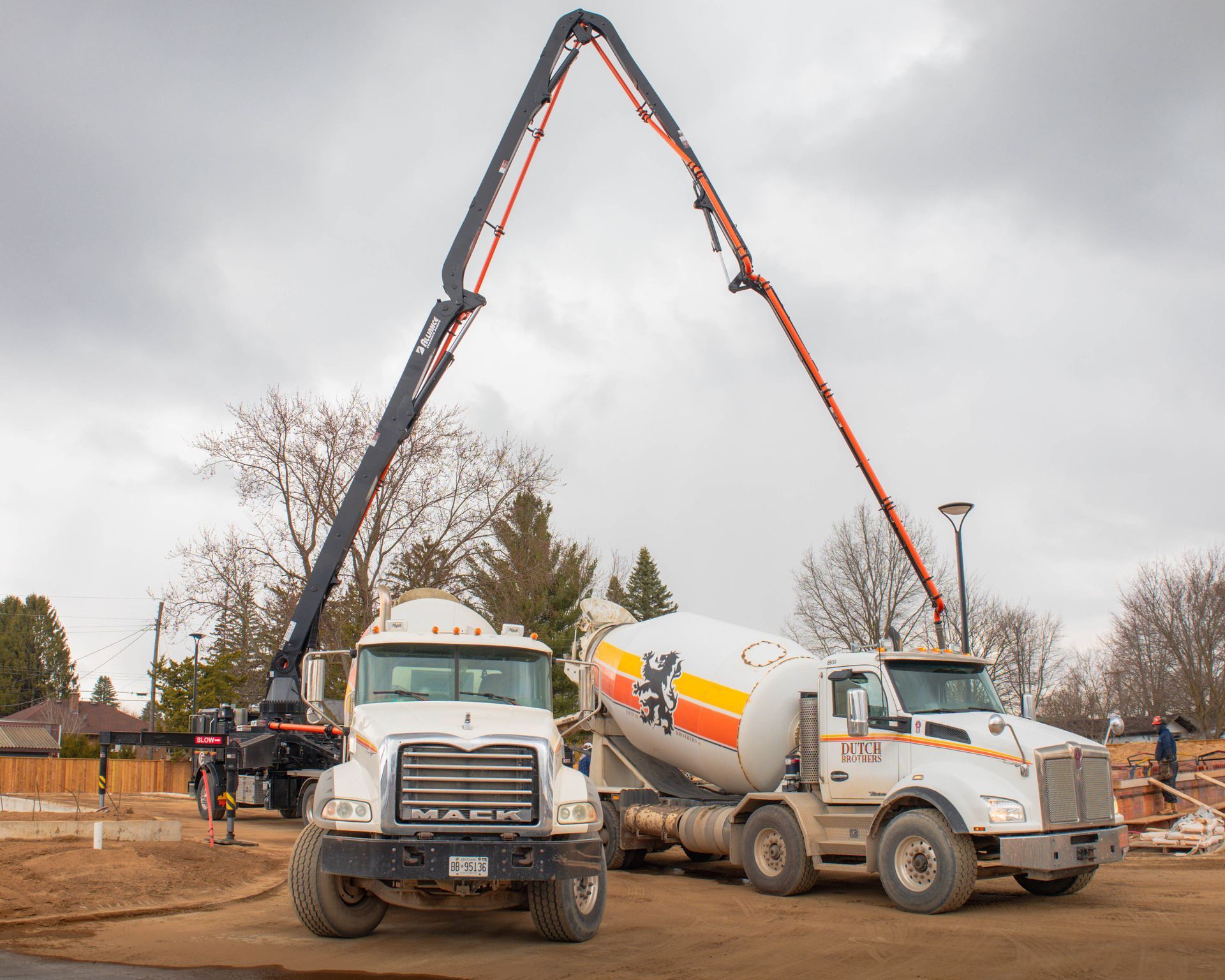 Two concrete trucks with an extended pump arm pouring concrete at a construction site under cloudy skies.