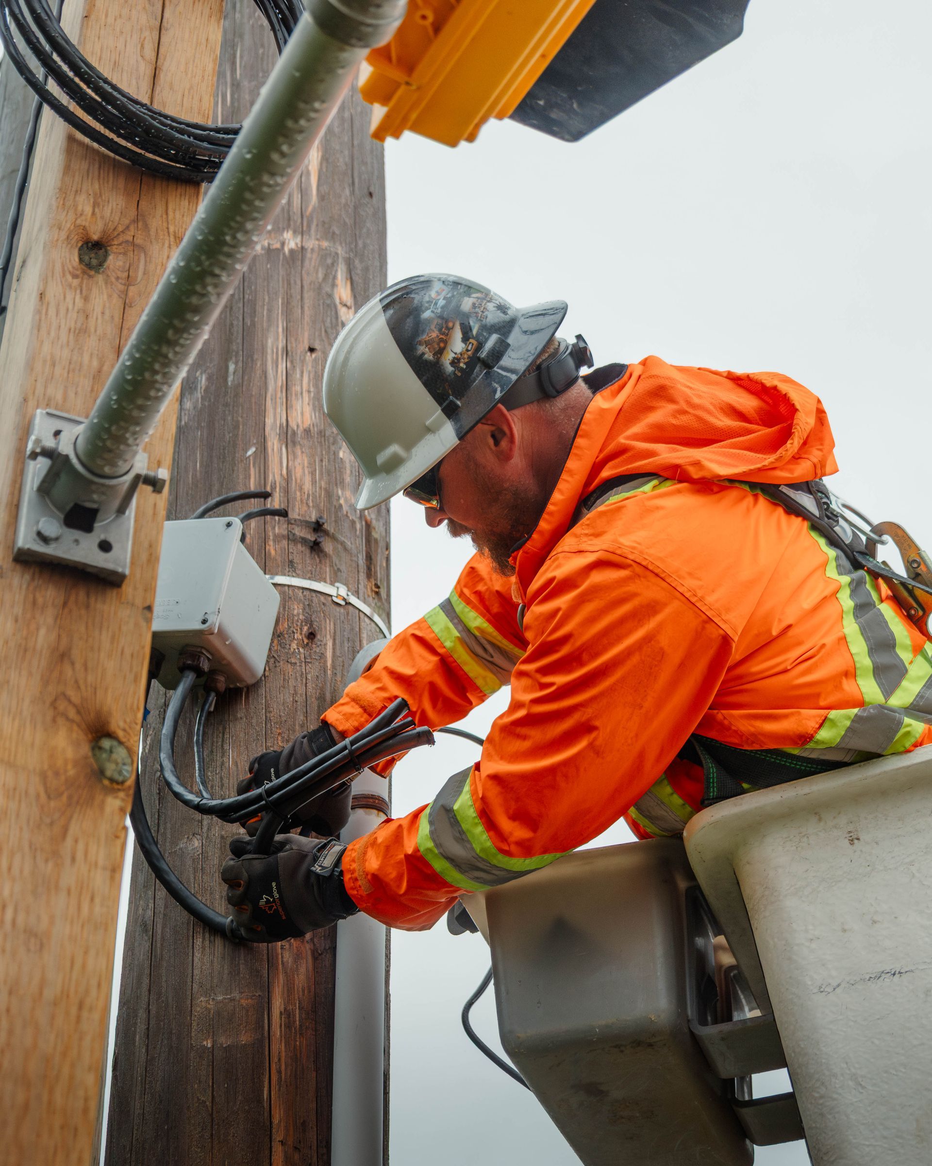 Lineman in orange jacket working on wires attached to a wooden utility pole.