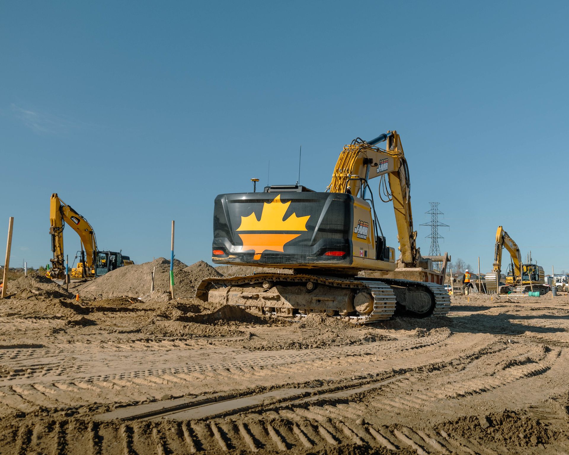 Construction site with three yellow excavators on dirt under a blue sky. One has a maple leaf design.