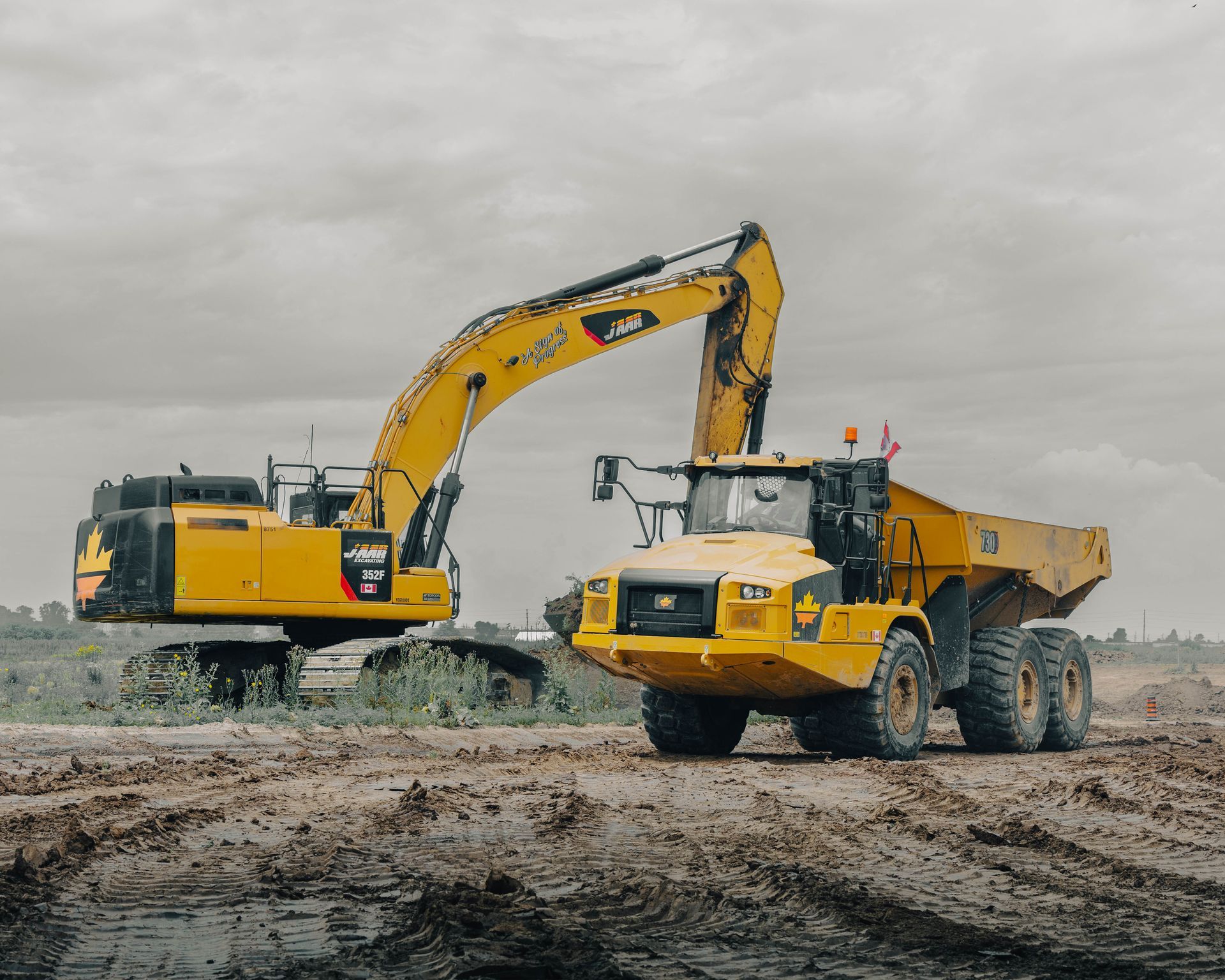 Yellow excavator loading a matching dump truck on a dirt worksite under a cloudy sky.