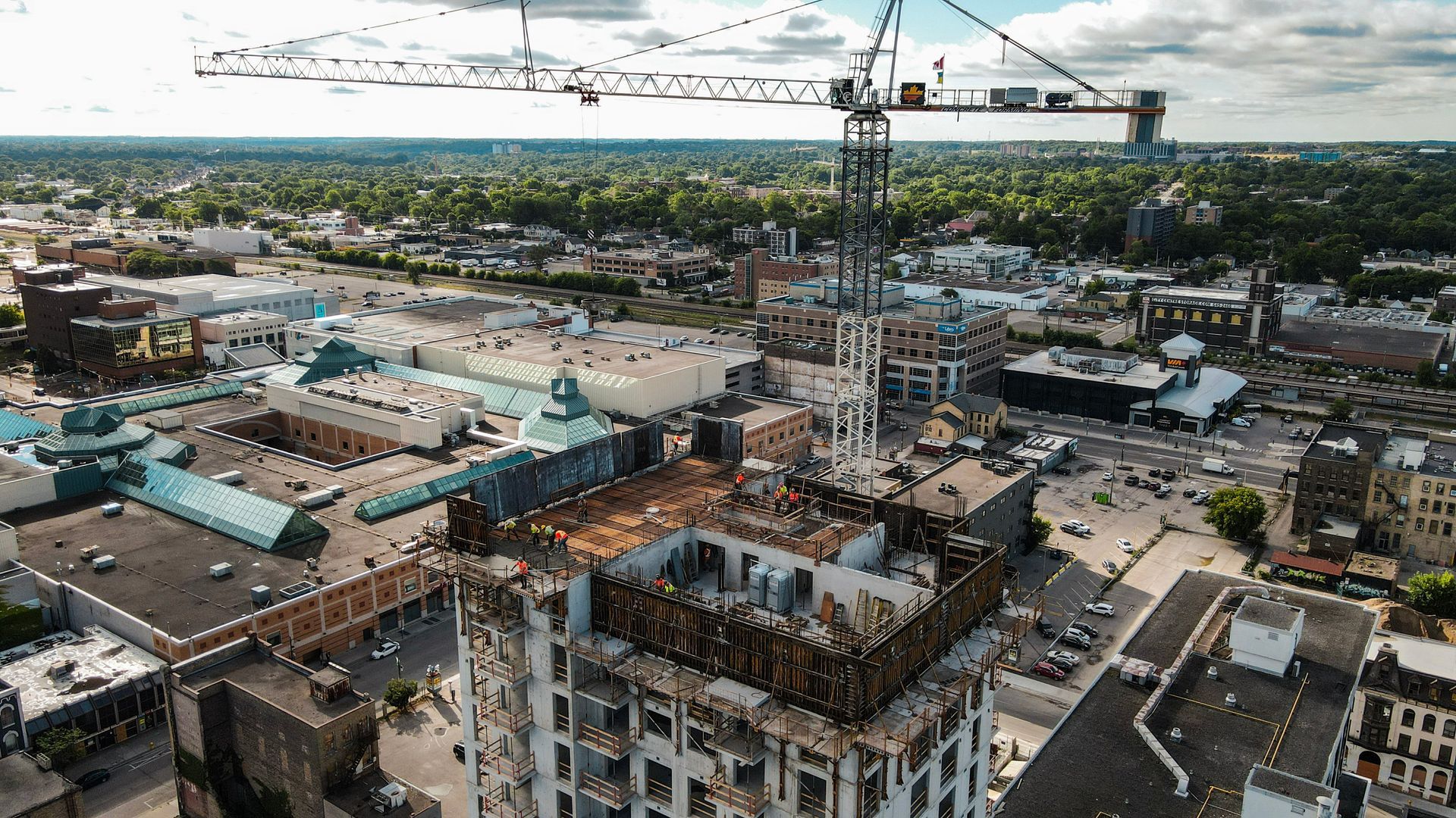 Overhead view of a city construction site with a large crane, buildings, and green trees in the background.