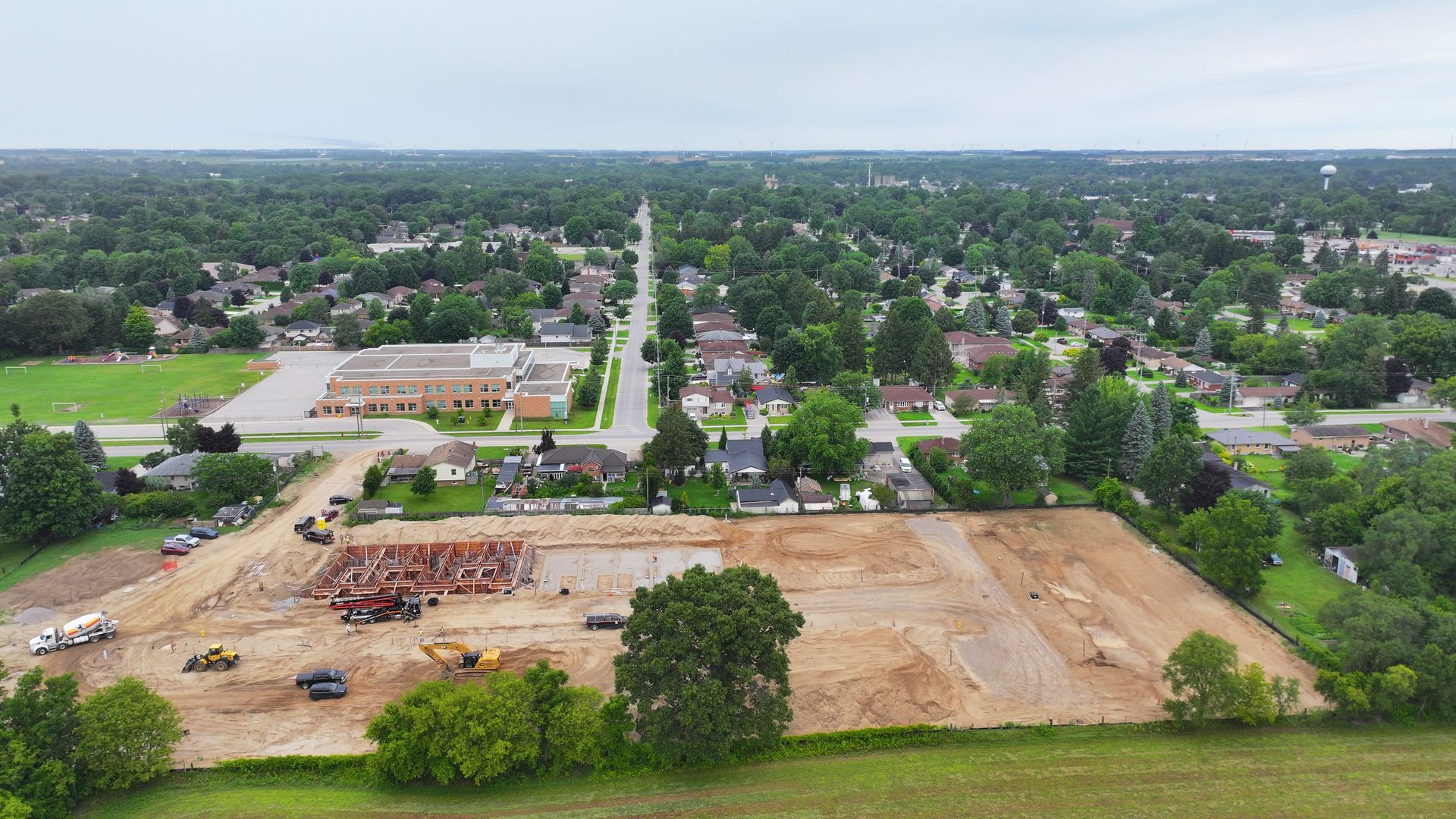 Aerial view of construction site with surrounding houses, trees, and road under overcast sky.