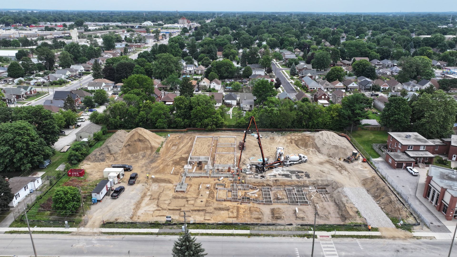 Aerial view of construction site. Foundation laid, crane, concrete truck, surrounded by dirt piles and residential area.
