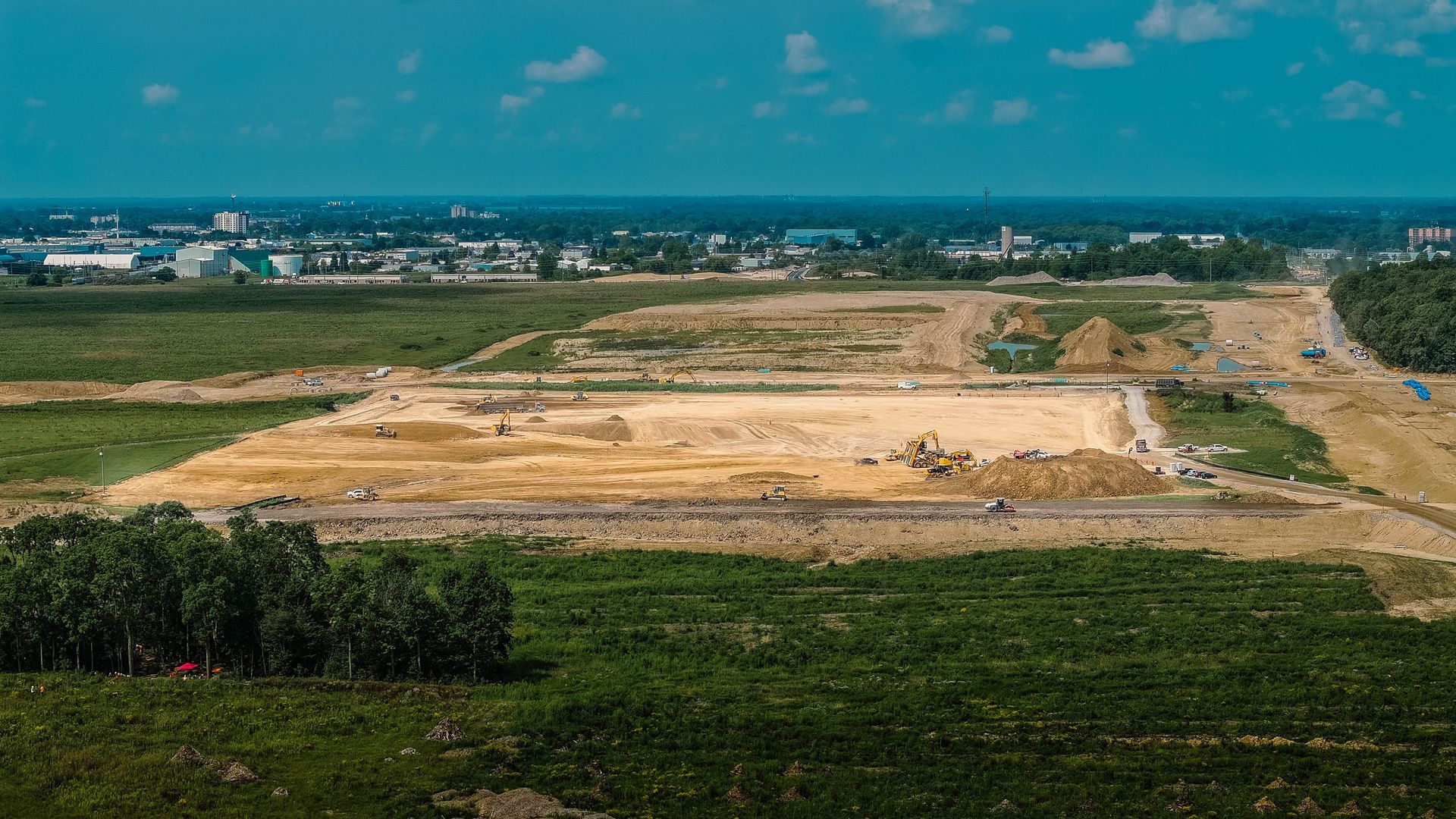 Construction site with cleared land, dirt piles, and a distant town under a blue sky.