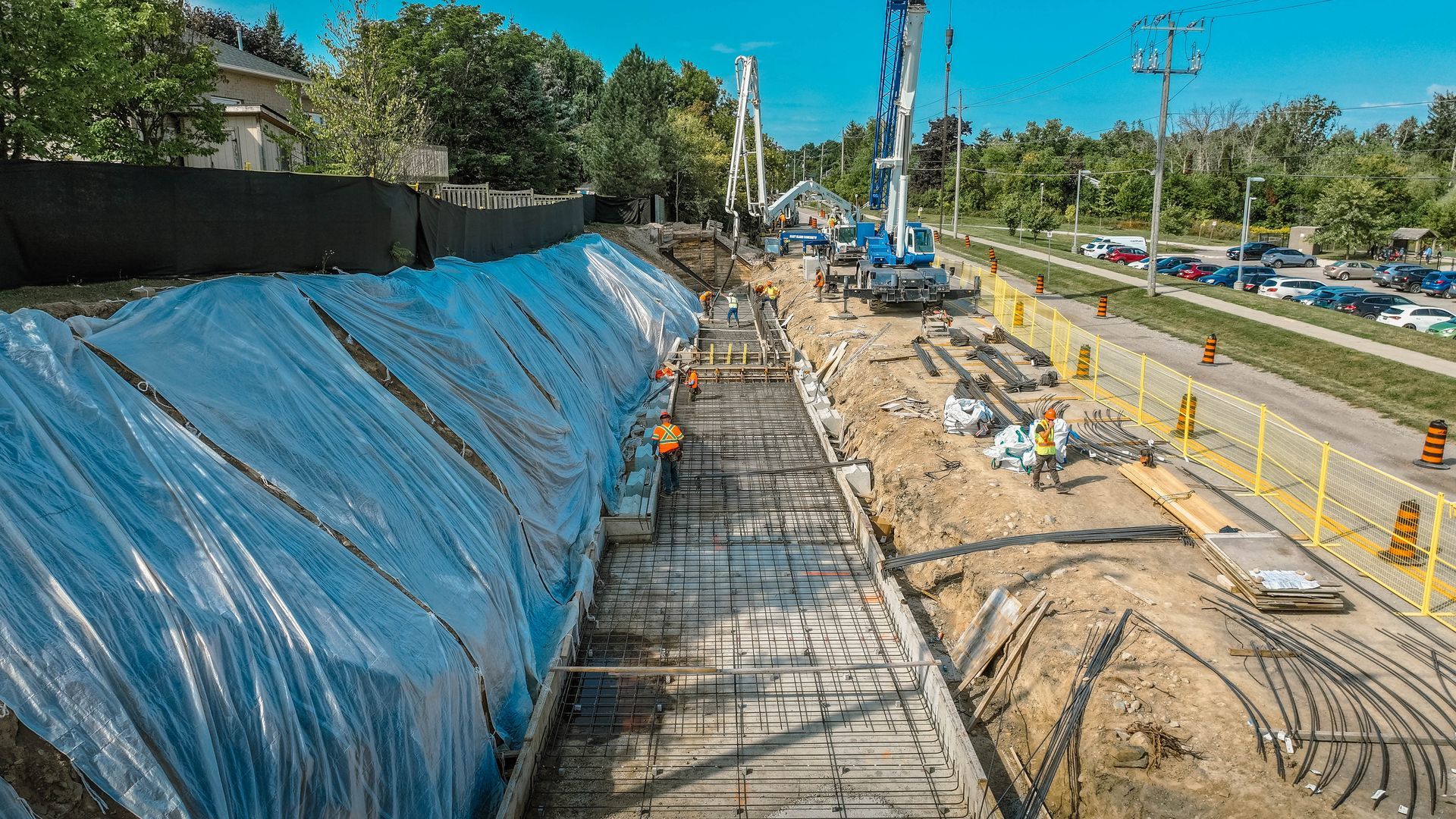 Construction site with workers, machinery, and concrete work. Blue tarp covers one side, road and trees in background.