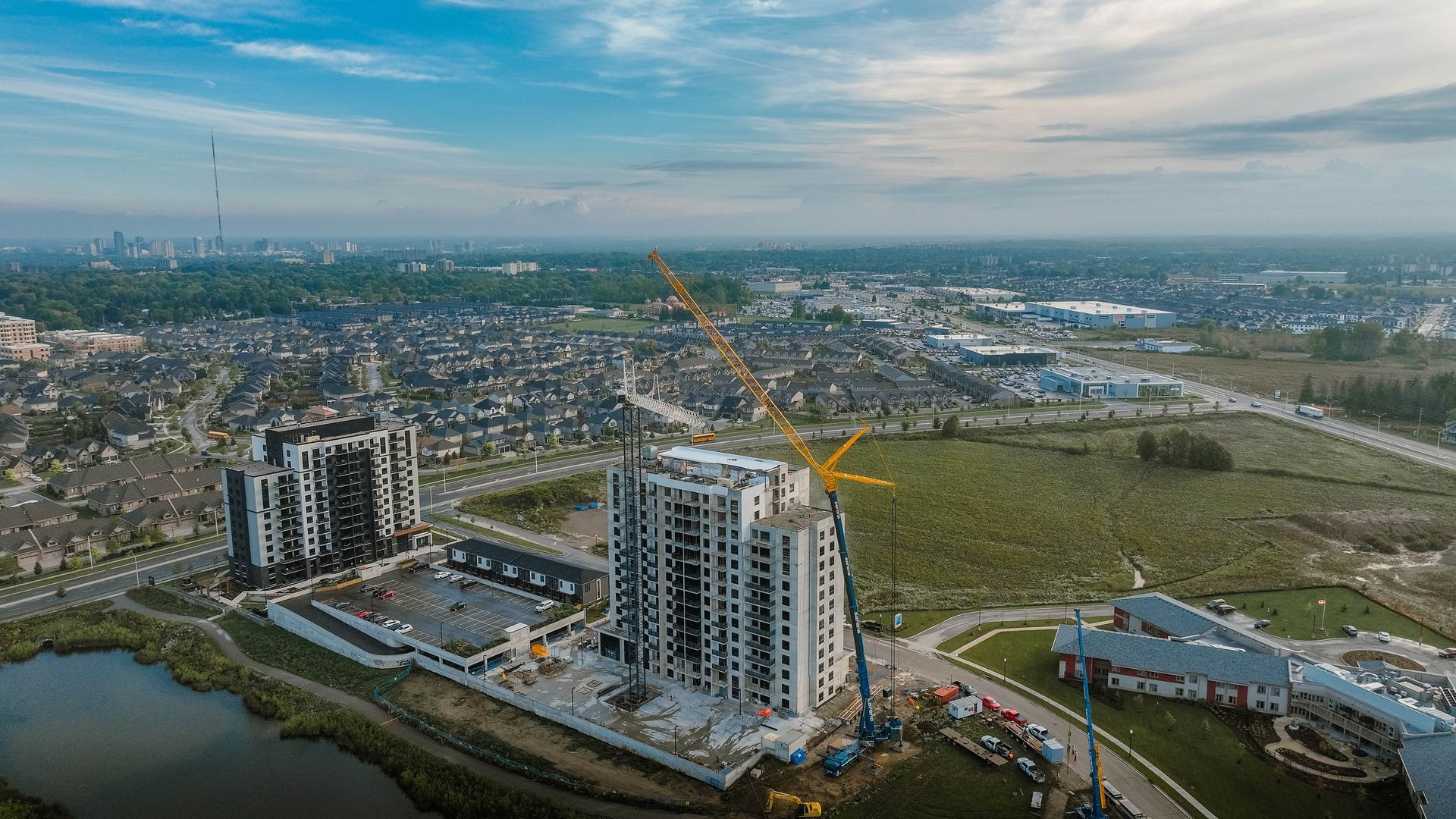 Aerial view of a city under construction; tall buildings, a crane, and a pond are visible under a cloudy sky.