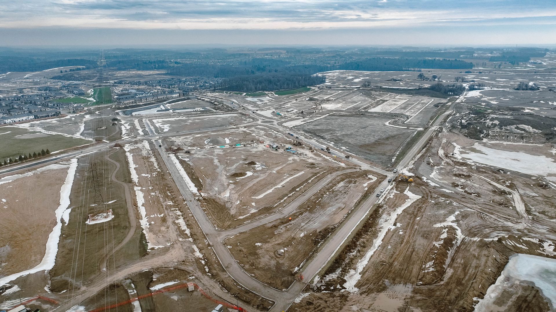 Aerial view of a construction site with roads and patches of snow, with a forest in the background.