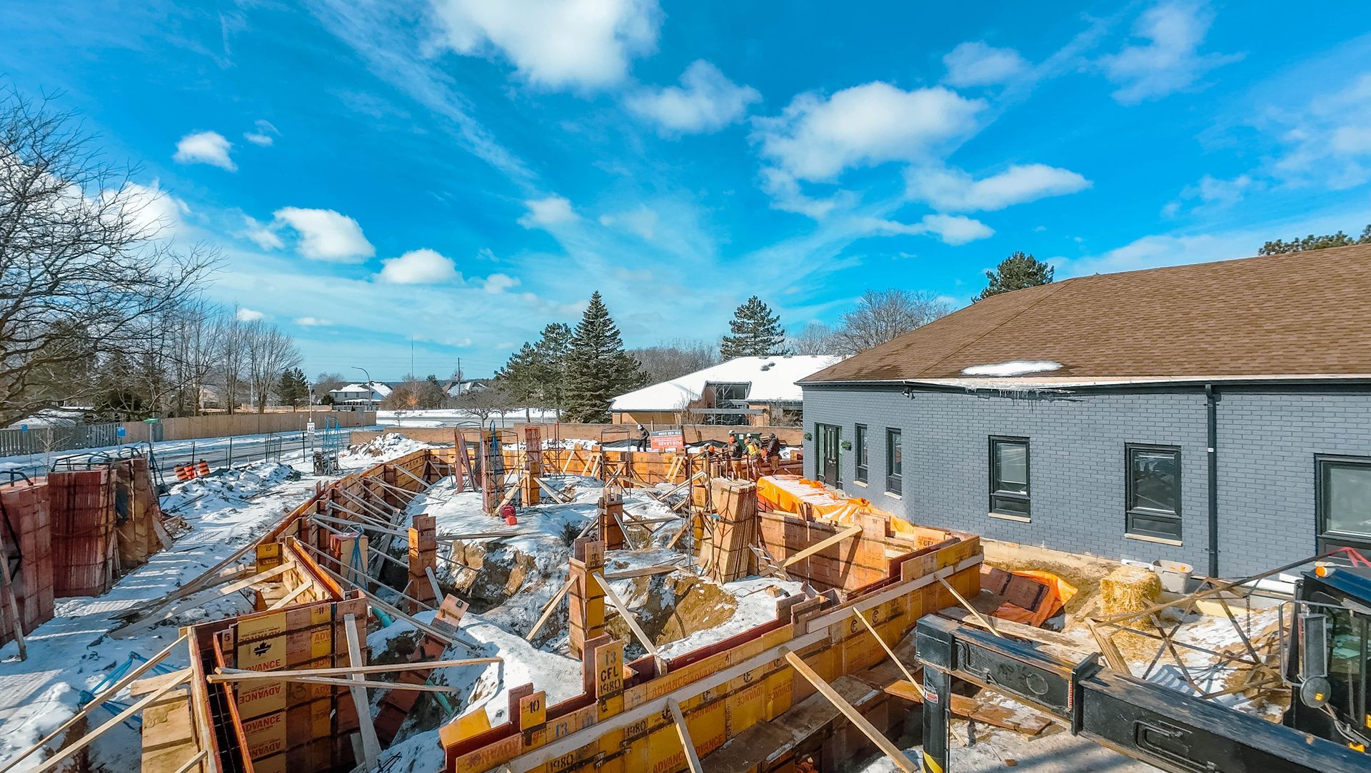 Construction site with wood forms, snow, blue sky, and a house under construction.