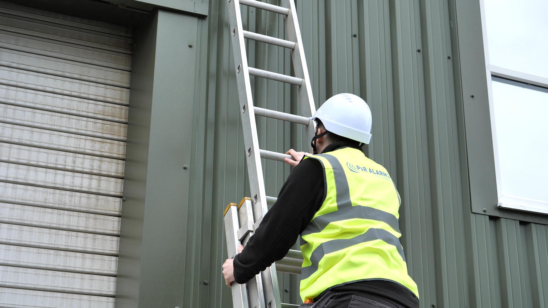 Man on ladder installing a security camera on a building exterior.