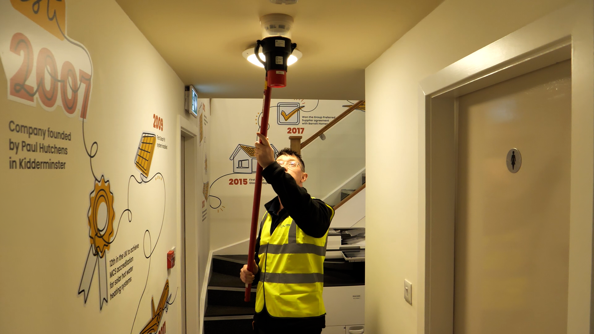 Person installing a ceiling-mounted device; wearing an orange vest and holding a tool. Light-colored room.