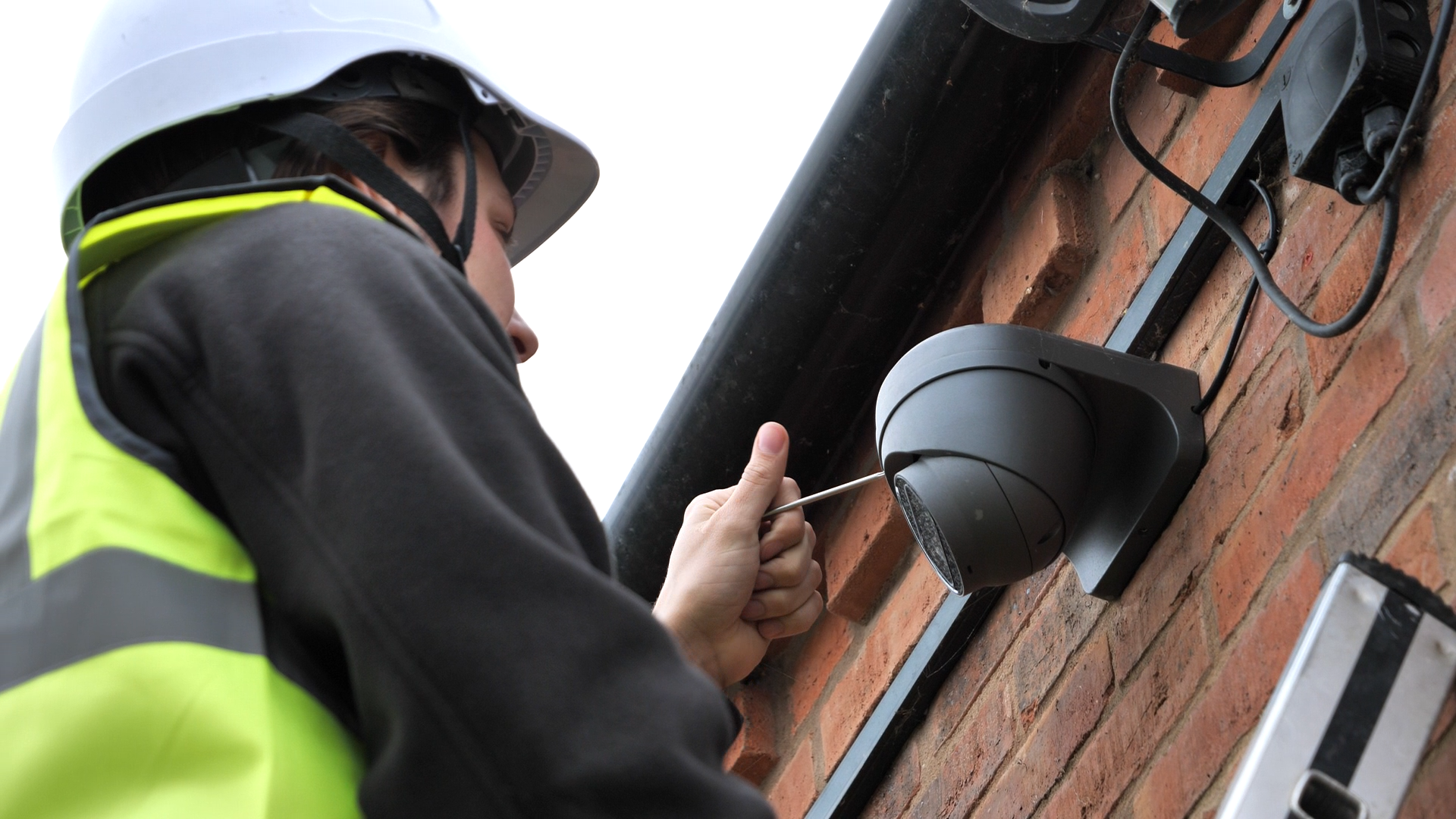 A person on a ladder installs a security camera on a building's exterior wall.